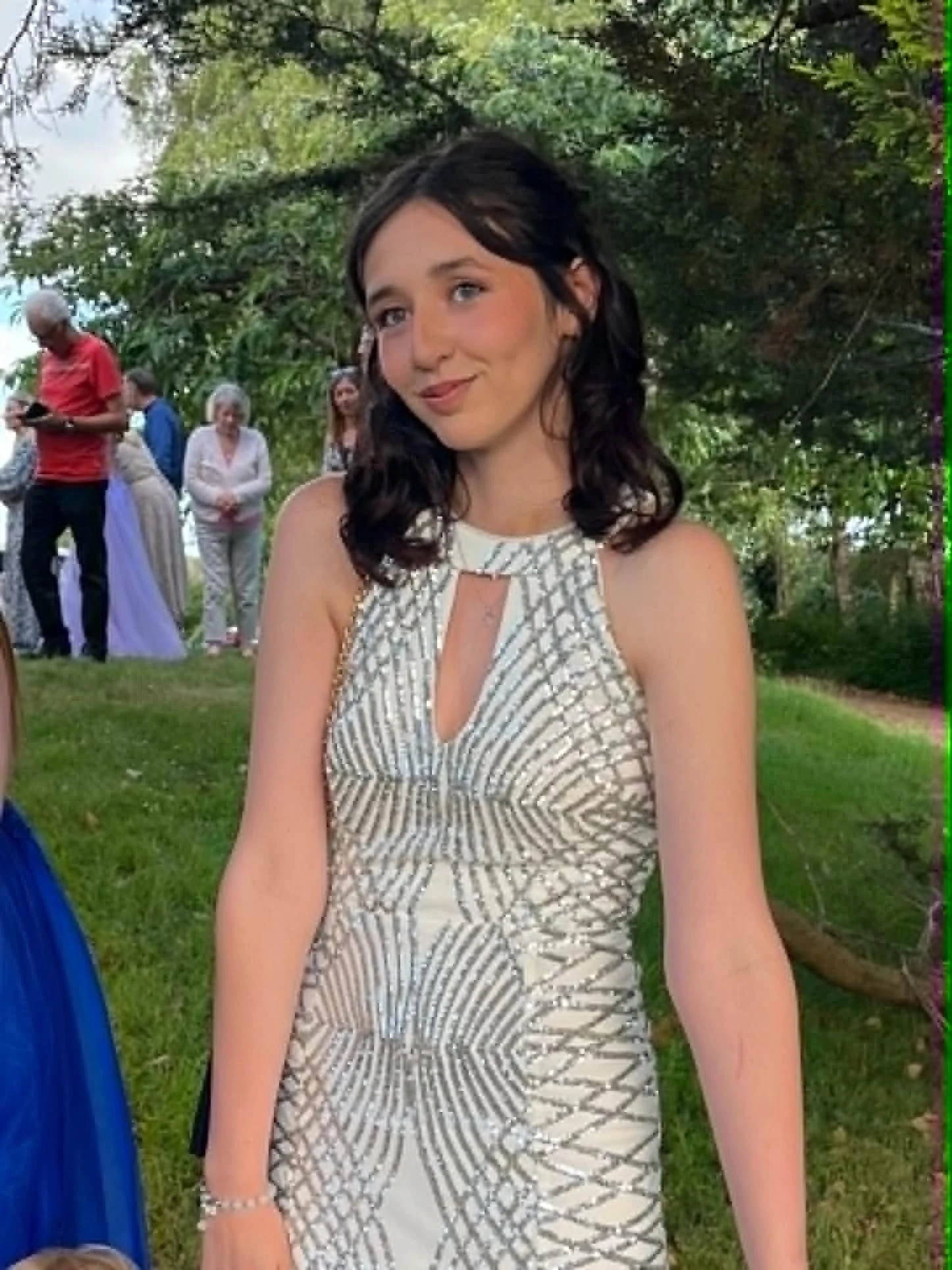 A young woman with dark wavy hair wearing a sleeveless, patterned dress with a keyhole neckline, smiling outdoors with green trees and several people in the background.