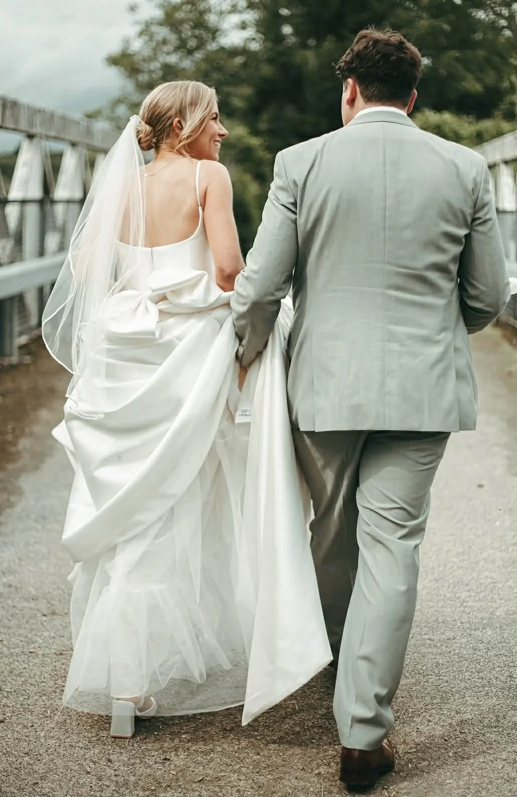 A bride and groom walking hand in hand outdoors on their wedding day, the bride holding up her wedding dress to reveal her shoes, with trees and a bridge in the background.