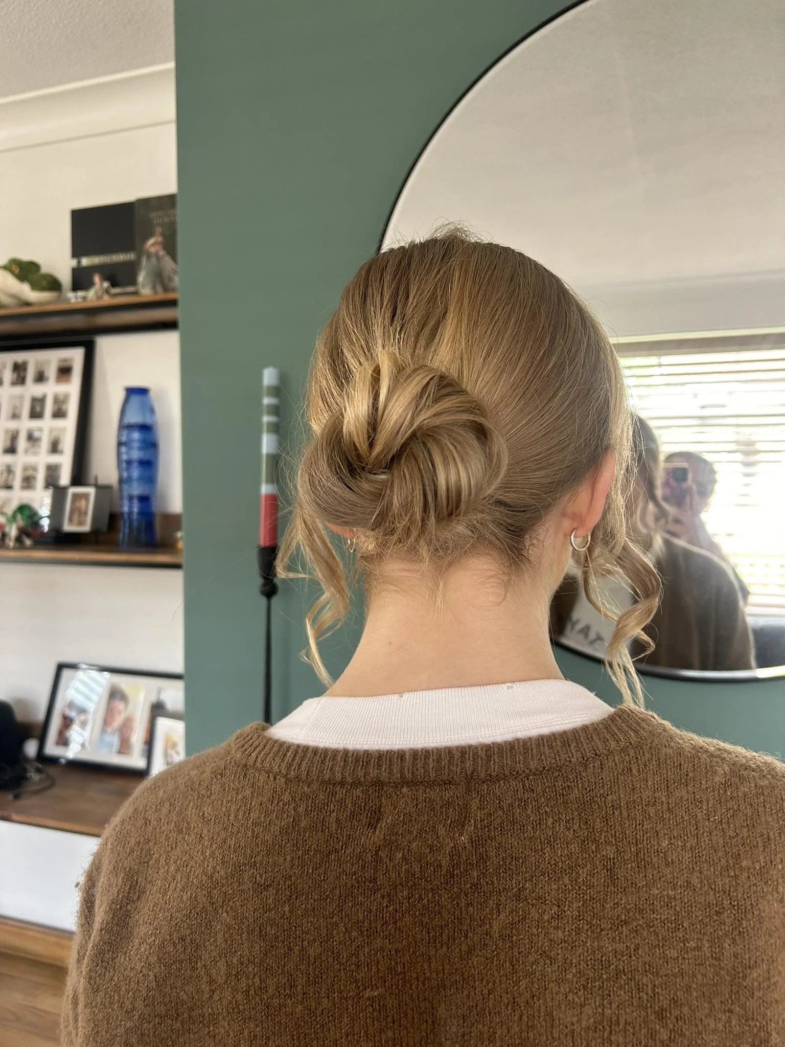 The back of a woman's head with styled blonde hair in a twisted bun, wearing a white shirt and brown sweater, reflected in a mirror in a room with various picture frames, a blue vase, and wall decor.