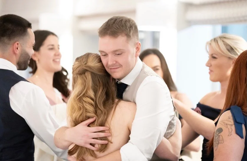 People dancing and hugging at a wedding reception, with a man and woman embracing in the center, others smiling around them.