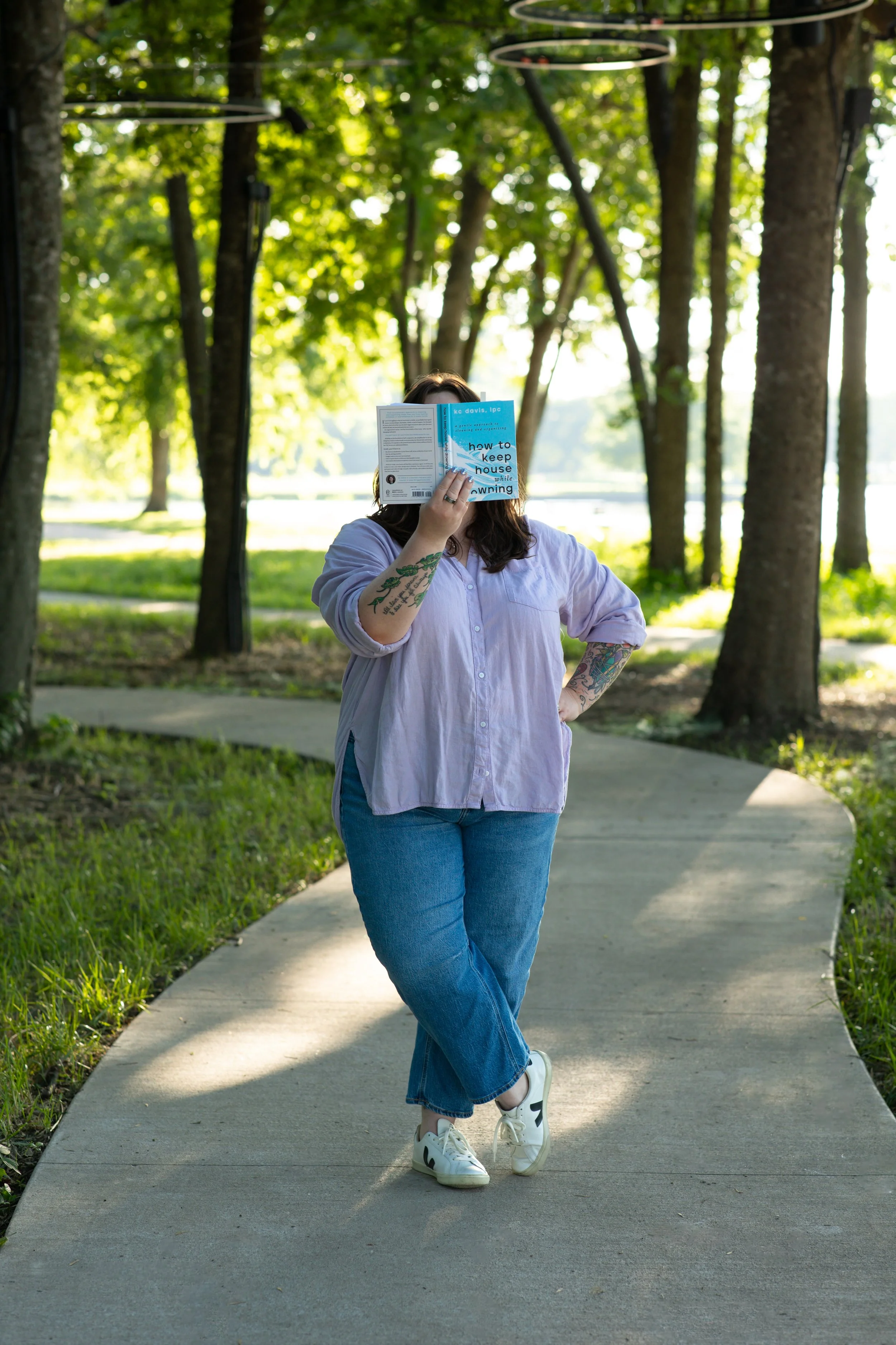 Person standing on a curved sidewalk in a park, holding a book in front of their face, wearing a lilac button-up shirt, blue jeans, and white sneakers, with tattoos visible on their arms, surrounded by trees with sunlight filtering through.