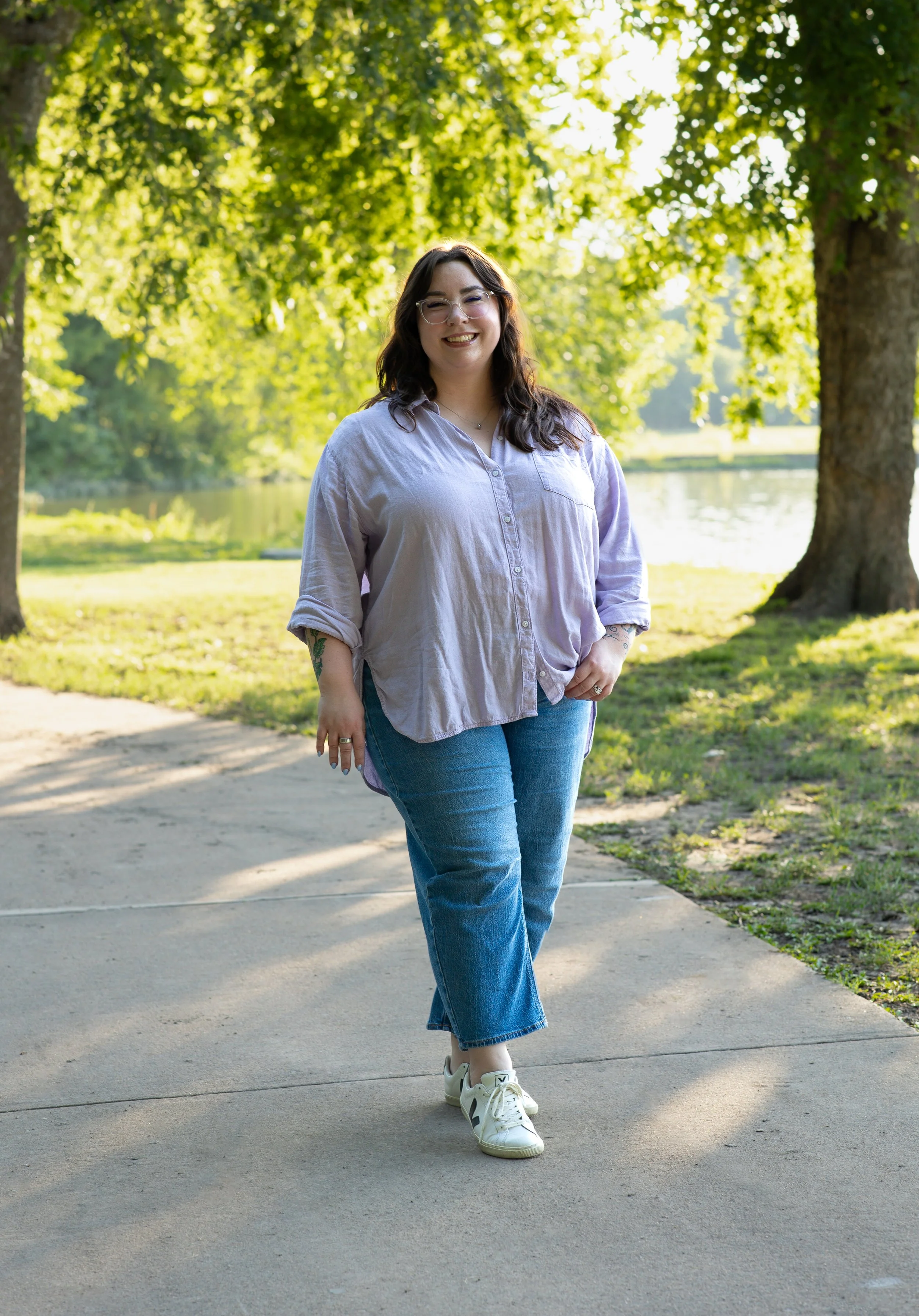 Woman walking outdoors in a park with trees and a river in the background, smiling at the camera, wearing glasses, a light purple shirt, jeans, and sneakers.