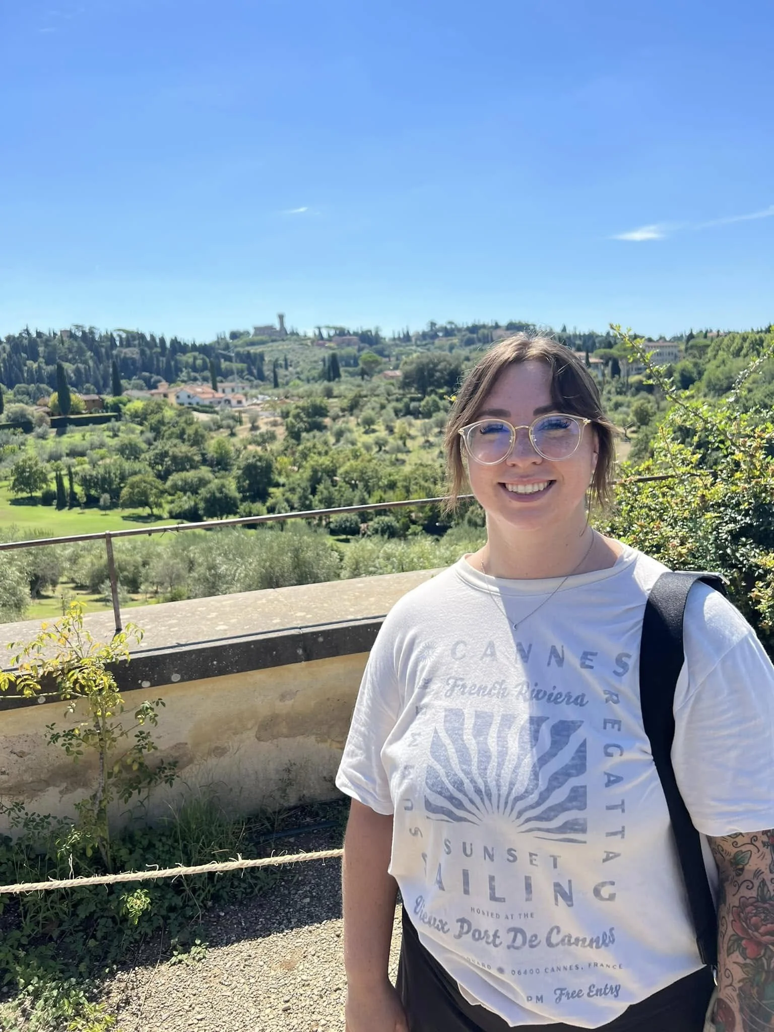 A woman smiling outdoors with a scenic hillside landscape of green trees, houses, and a distant tower under a blue sky in the background.