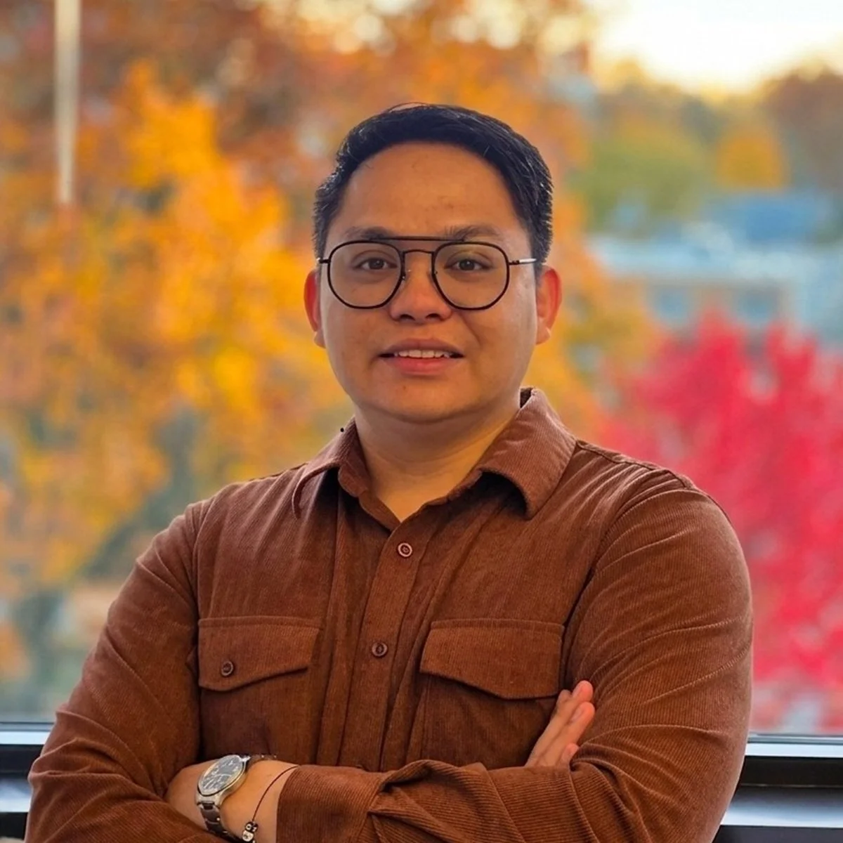 A man with glasses and short dark hair posing with arms crossed in front of a window with colorful autumn trees in the background.