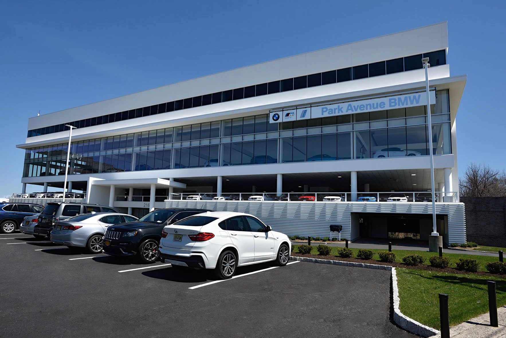 Modern four-story BMW dealership building with glass windows, displaying cars inside and in the parking lot, under a clear blue sky.