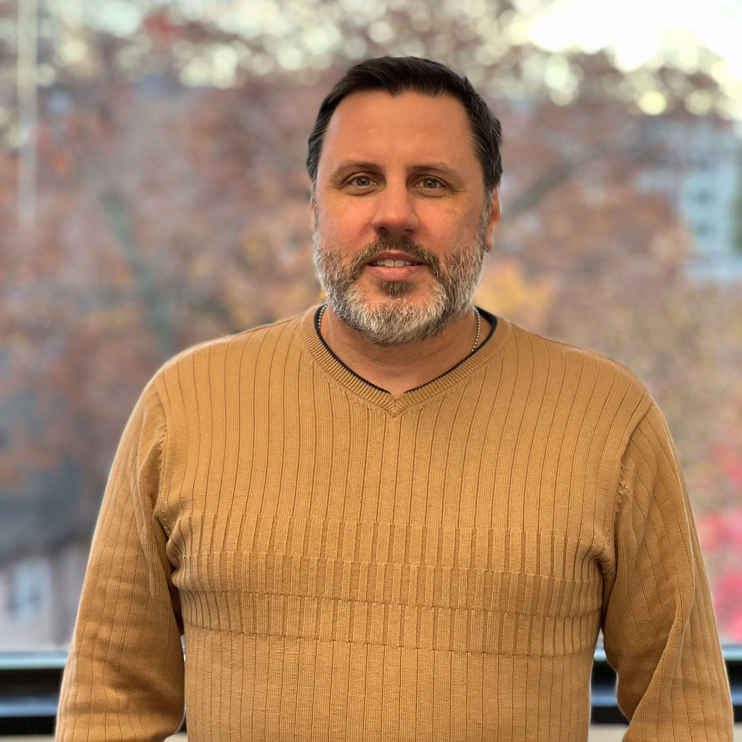 The image shows a close-up of a middle-aged man with dark hair and a beard, smiling at the camera. He is wearing a light brown, ribbed sweater. The background is blurred with colors suggesting an outdoor scene with trees and possibly some buildings.