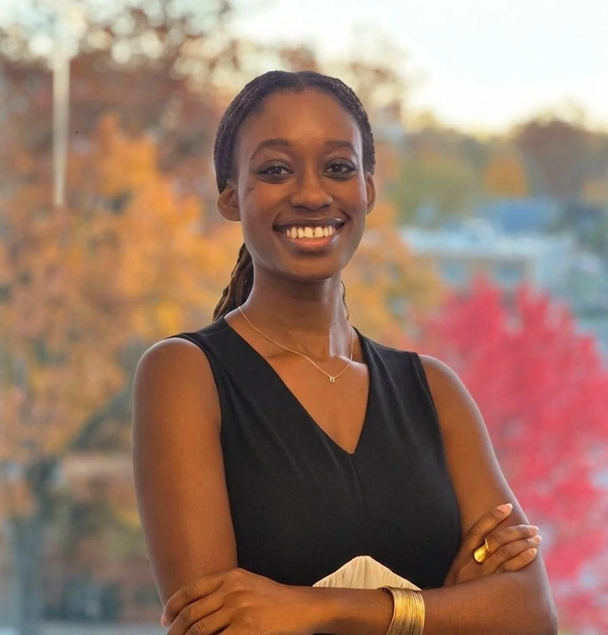 A young woman smiling with arms crossed, wearing a black sleeveless top, standing outdoors with autumn trees in the background.