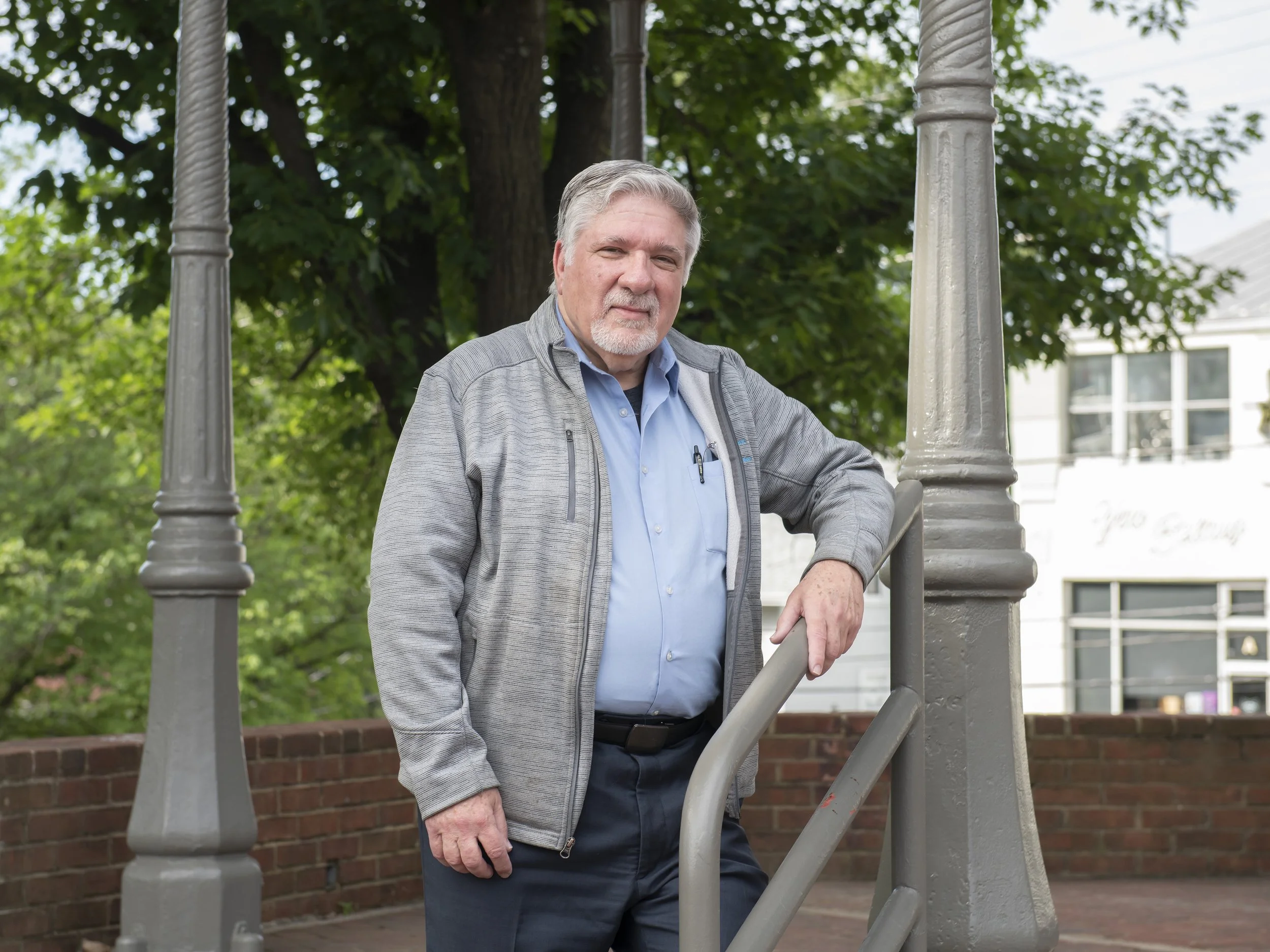 An older man with gray hair and a beard wearing a light blue shirt and gray jacket standing outdoors on a staircase with green trees and buildings in the background.