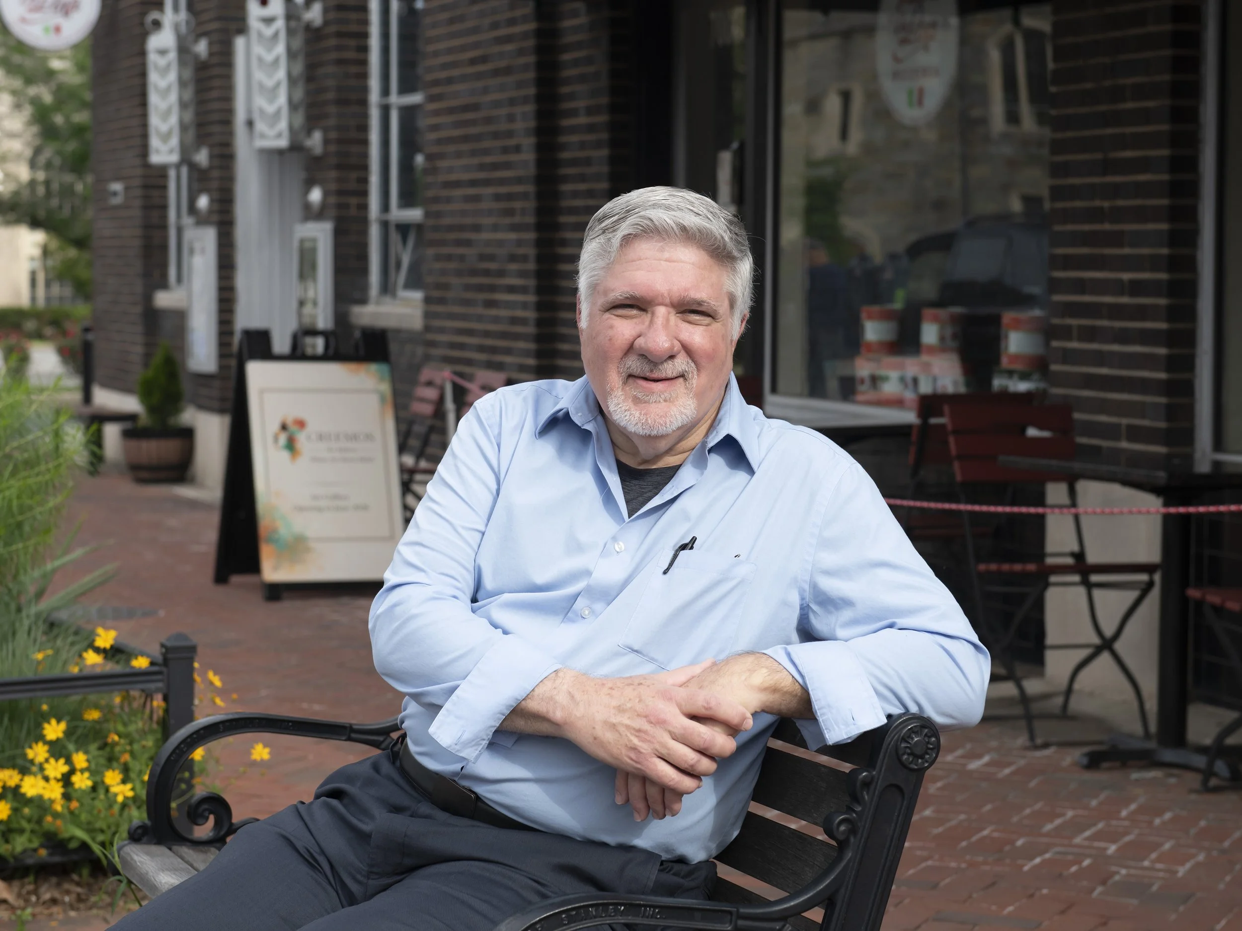 A smiling older man with gray hair and a beard, wearing a light blue shirt, sitting on a bench outside a brick building with large windows, surrounded by yellow flowers and greenery.