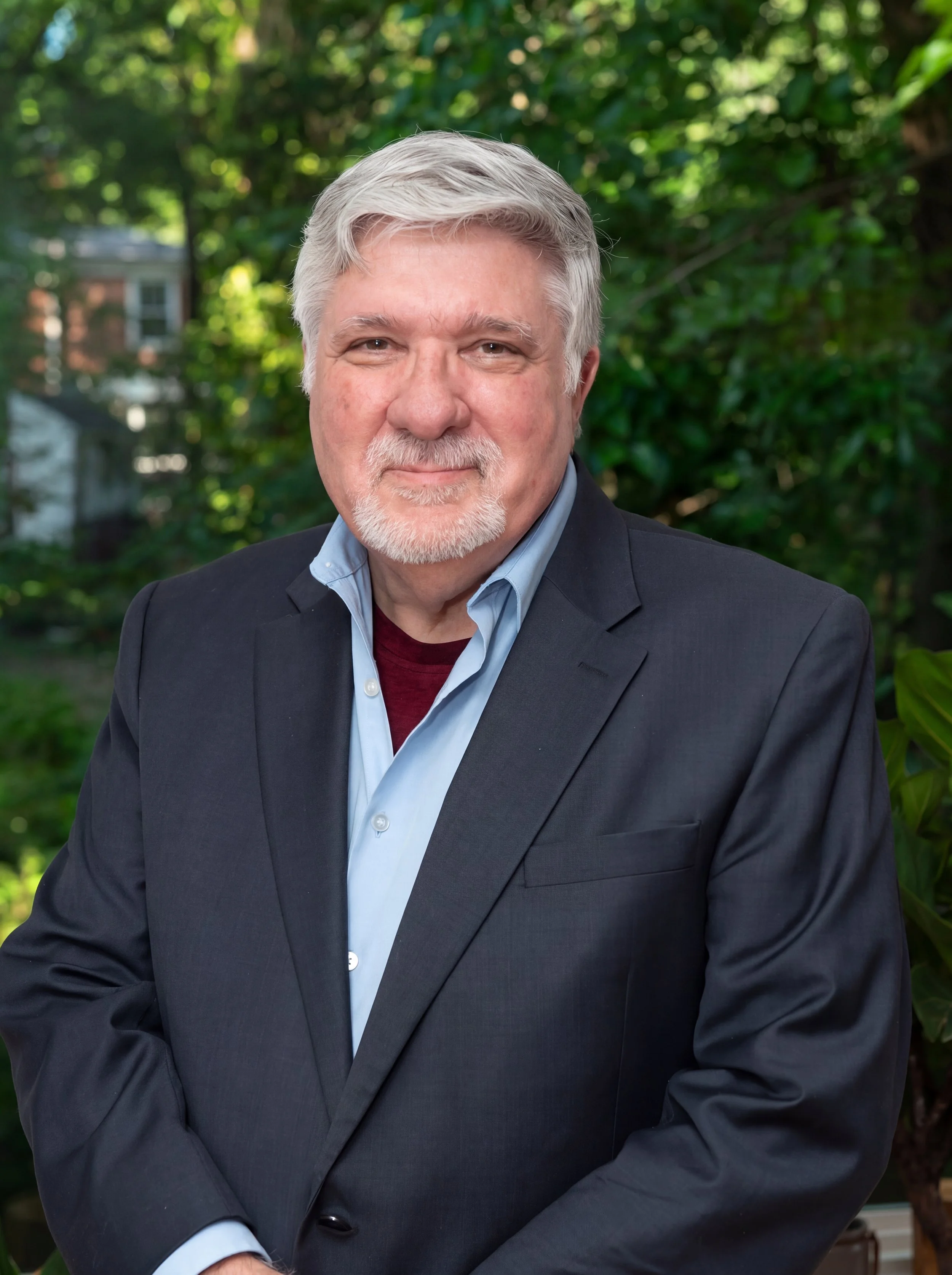 A mature man with white hair and a beard, wearing a navy blazer and light blue shirt, standing outdoors with green trees and buildings in the background.