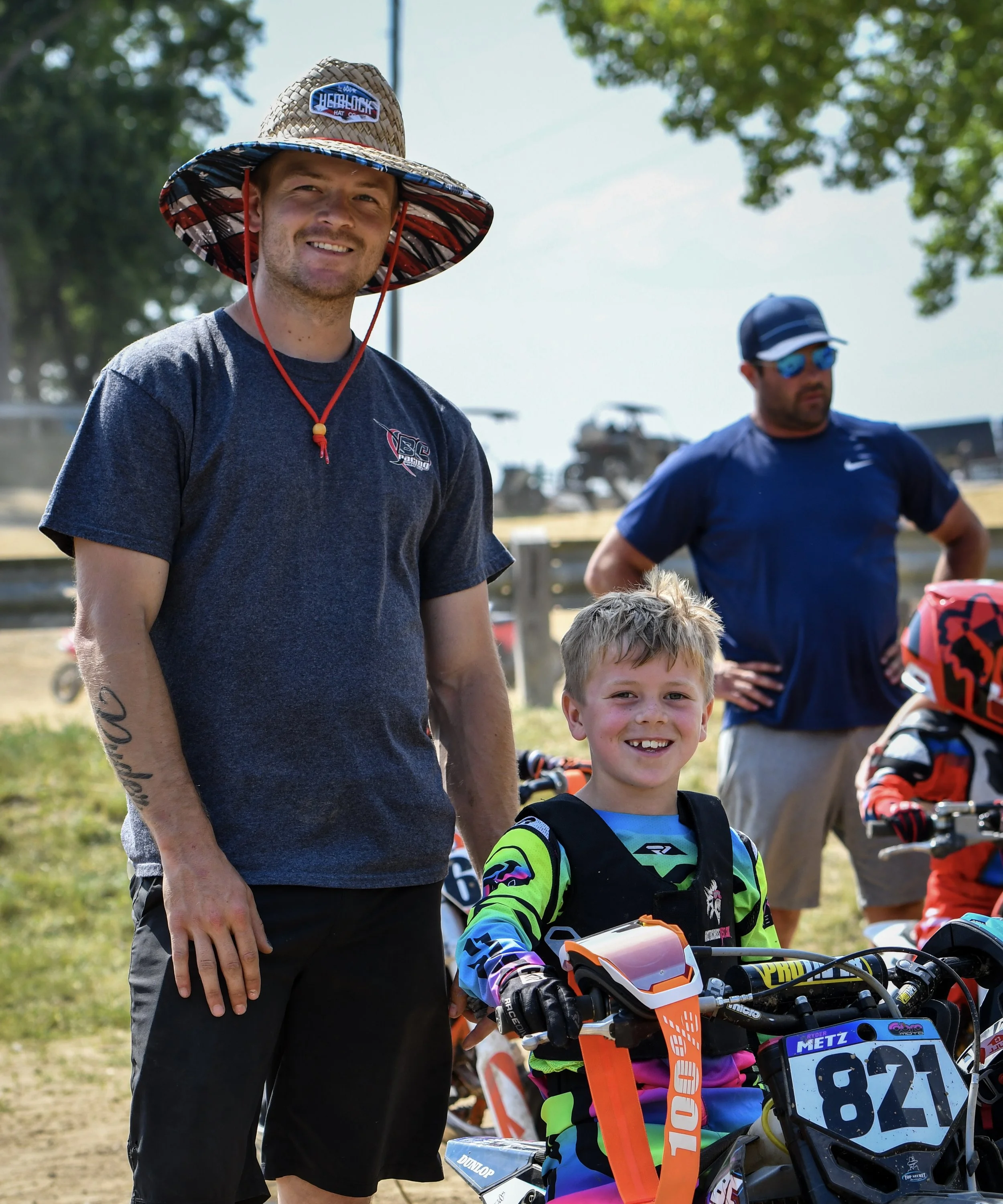 A smiling man wearing a straw hat with a patch and a gray t-shirt stands next to a young boy in colorful motocross gear and helmet, both outdoors on a sunny day.