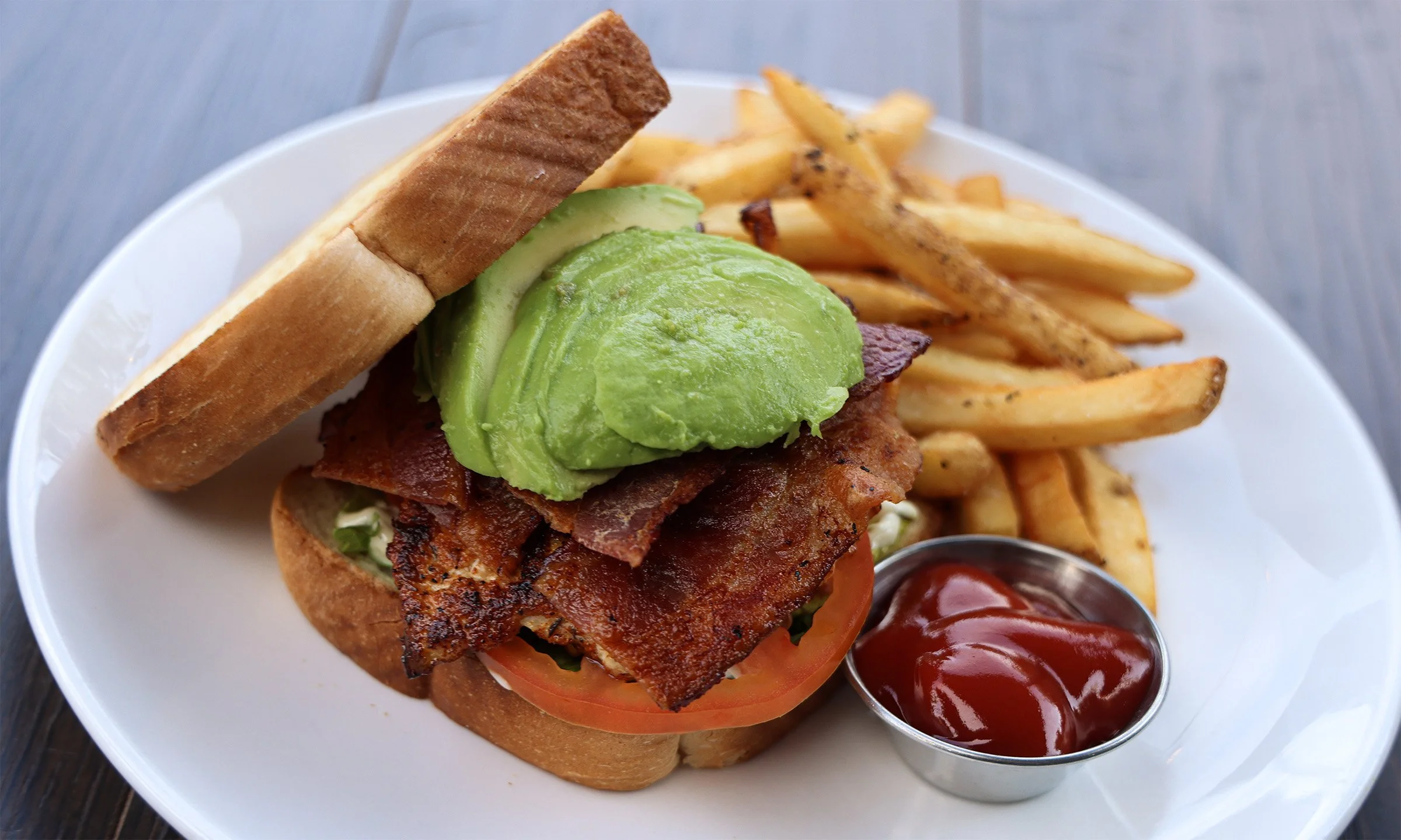 A plate with a sandwich containing bacon, tomato, lettuce, avocado, and bread, served with French fries and ketchup.