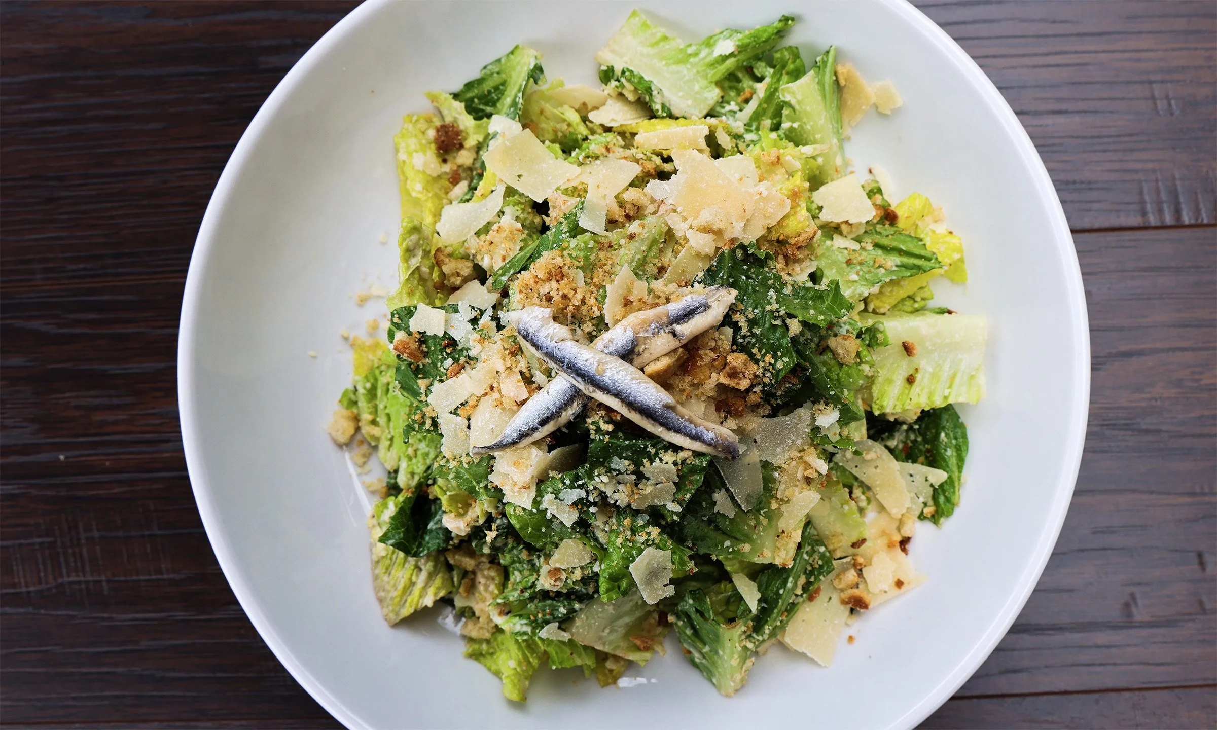Mixed green salad with anchovies, cheese shavings, and croutons on a white plate