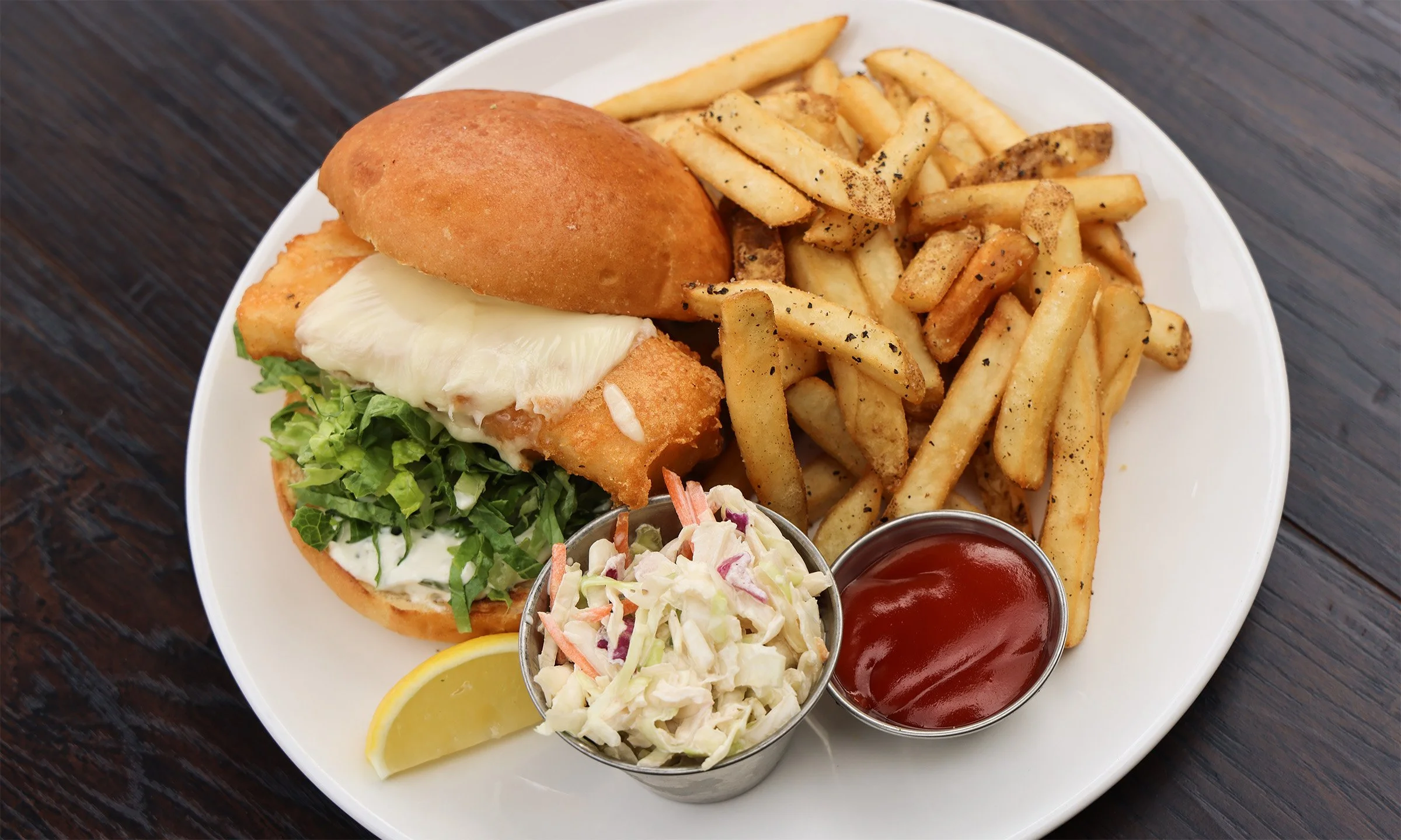 A plate of fish sandwich with French fries, coleslaw, ketchup, and a lemon wedge