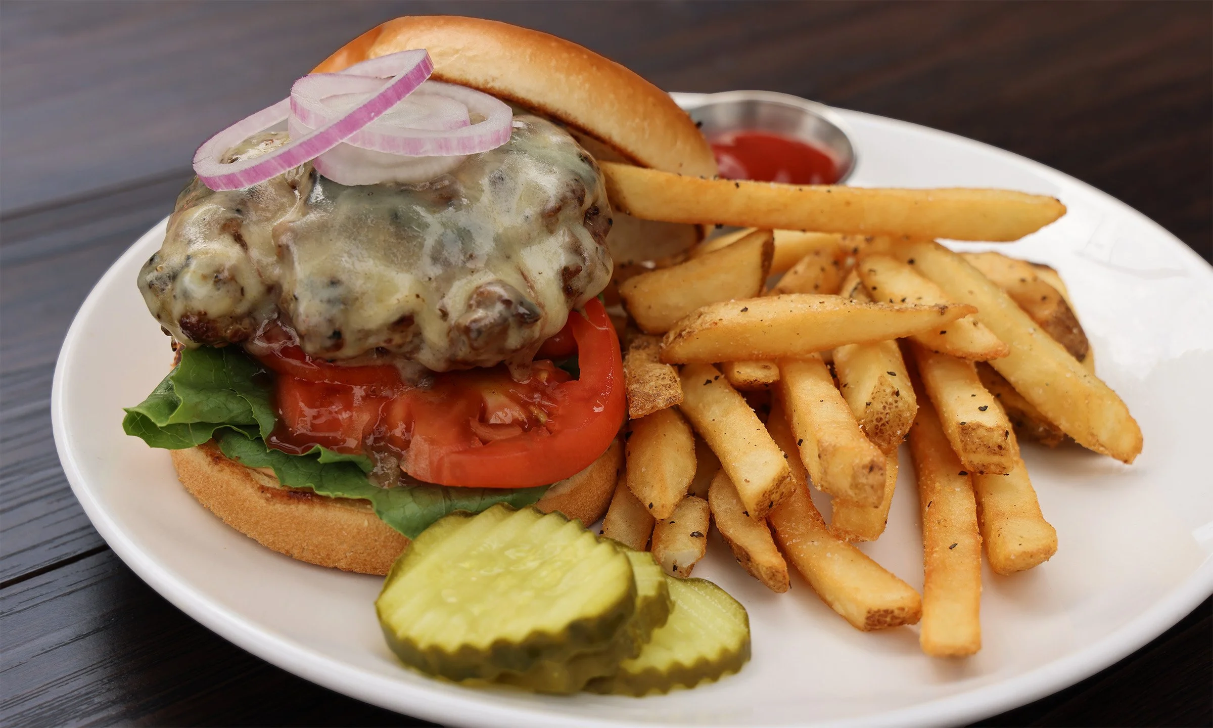 Plate with cheeseburger topped with onions, lettuce, tomato, pickles, and a bun, served with French fries and ketchup