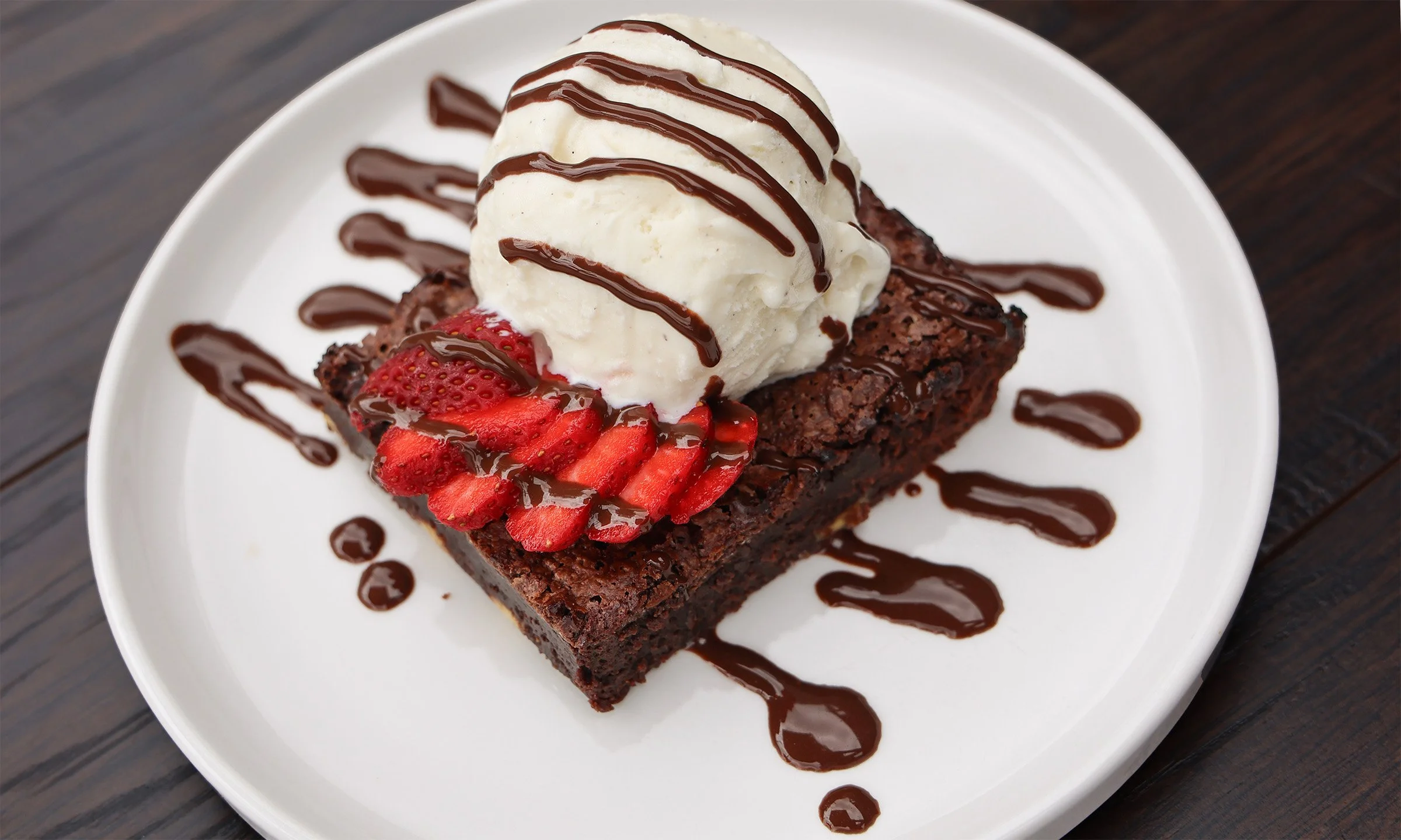 Chocolate brownie topped with vanilla ice cream, sliced strawberries, and drizzled with chocolate sauce on a white plate