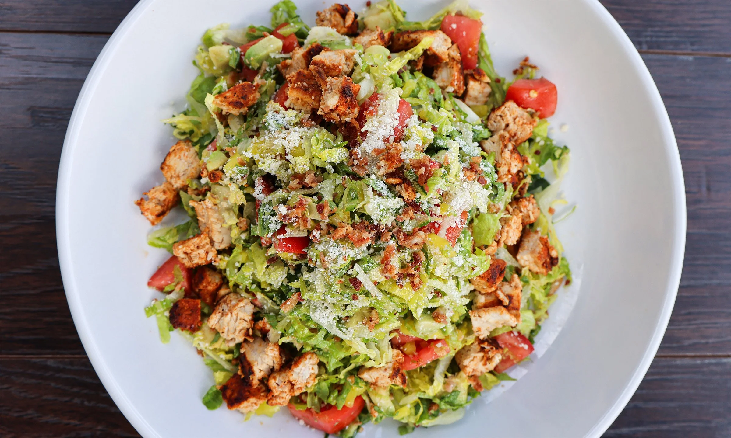 Salad with chopped lettuce, grilled chicken pieces, diced tomatoes, shredded cheese, and croutons in a white bowl on a wooden surface.