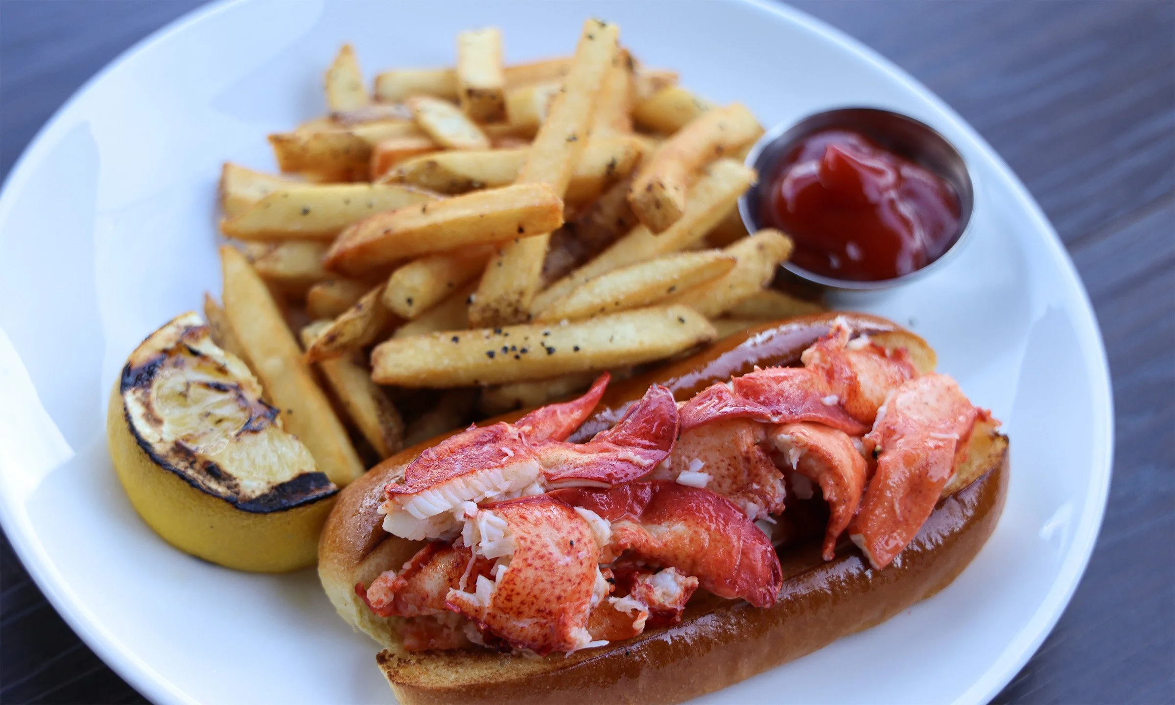 Lobster roll with fries, grilled lemon, and ketchup on a white plate.