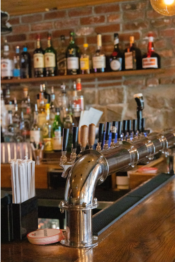 A bar counter with beer taps, straw holders, and liquor bottles on shelves against a brick wall in a cozy bar setting.