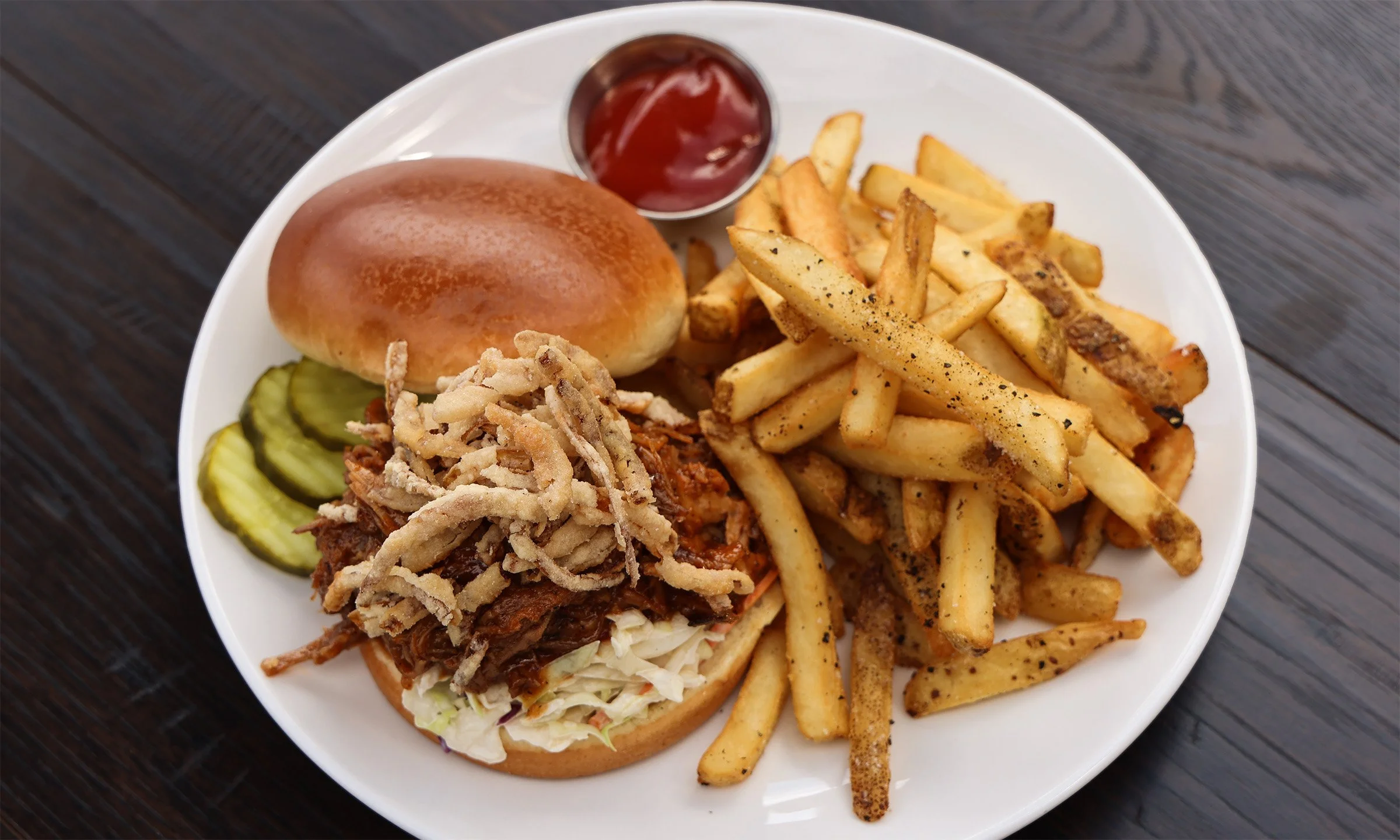 Barbecue pulled pork sandwich with fried onion strings, pickles, and a side of French fries, served with ketchup on a white plate.