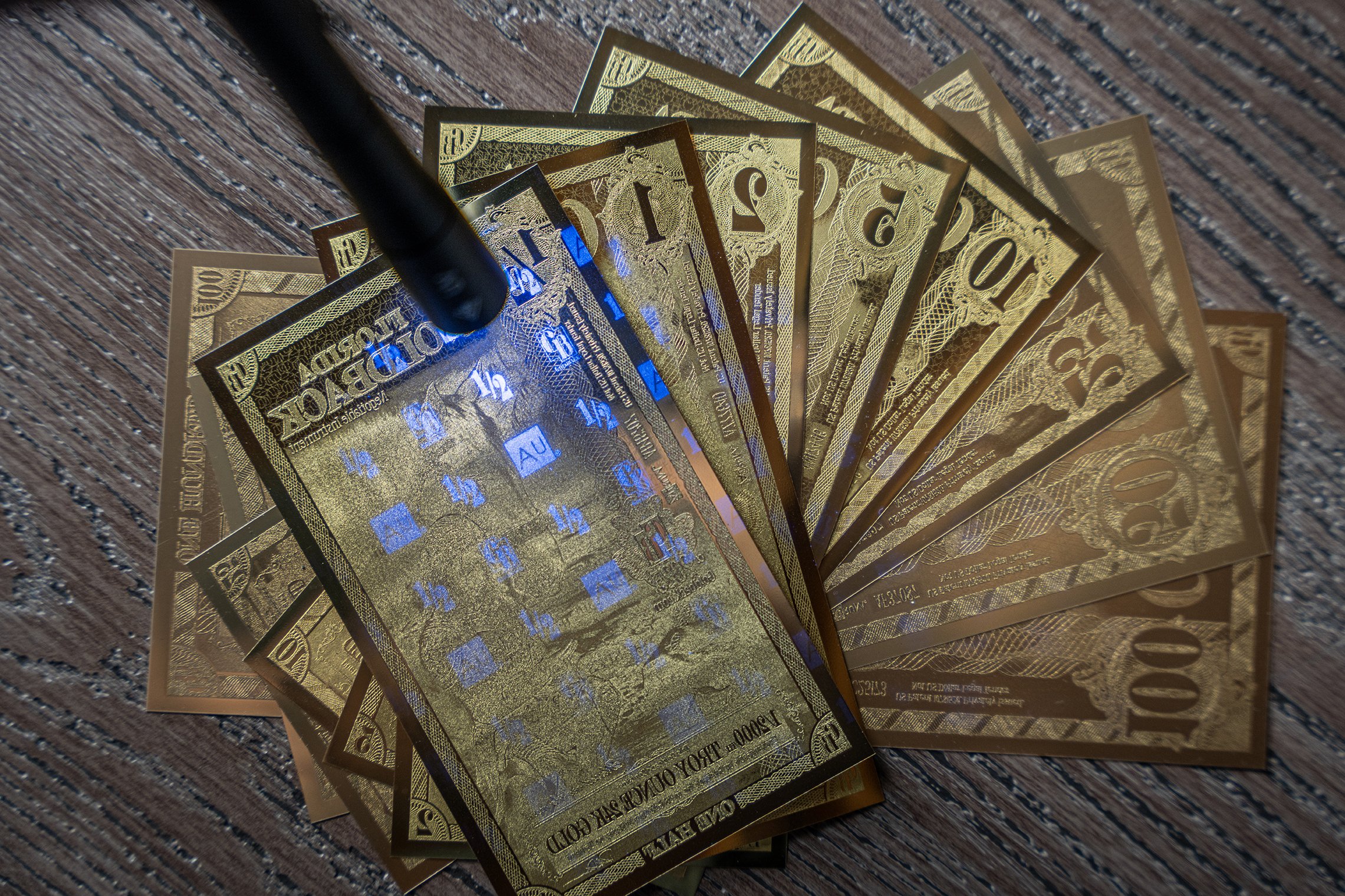 A fan of ten gold-colored English currency banknotes on a wooden surface with a black object partially covering the notes.