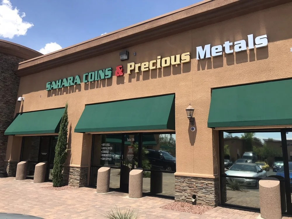 Exterior of a store called Sahara Coins & Precious Metals with green awnings and large glass windows, with parking lot in front.