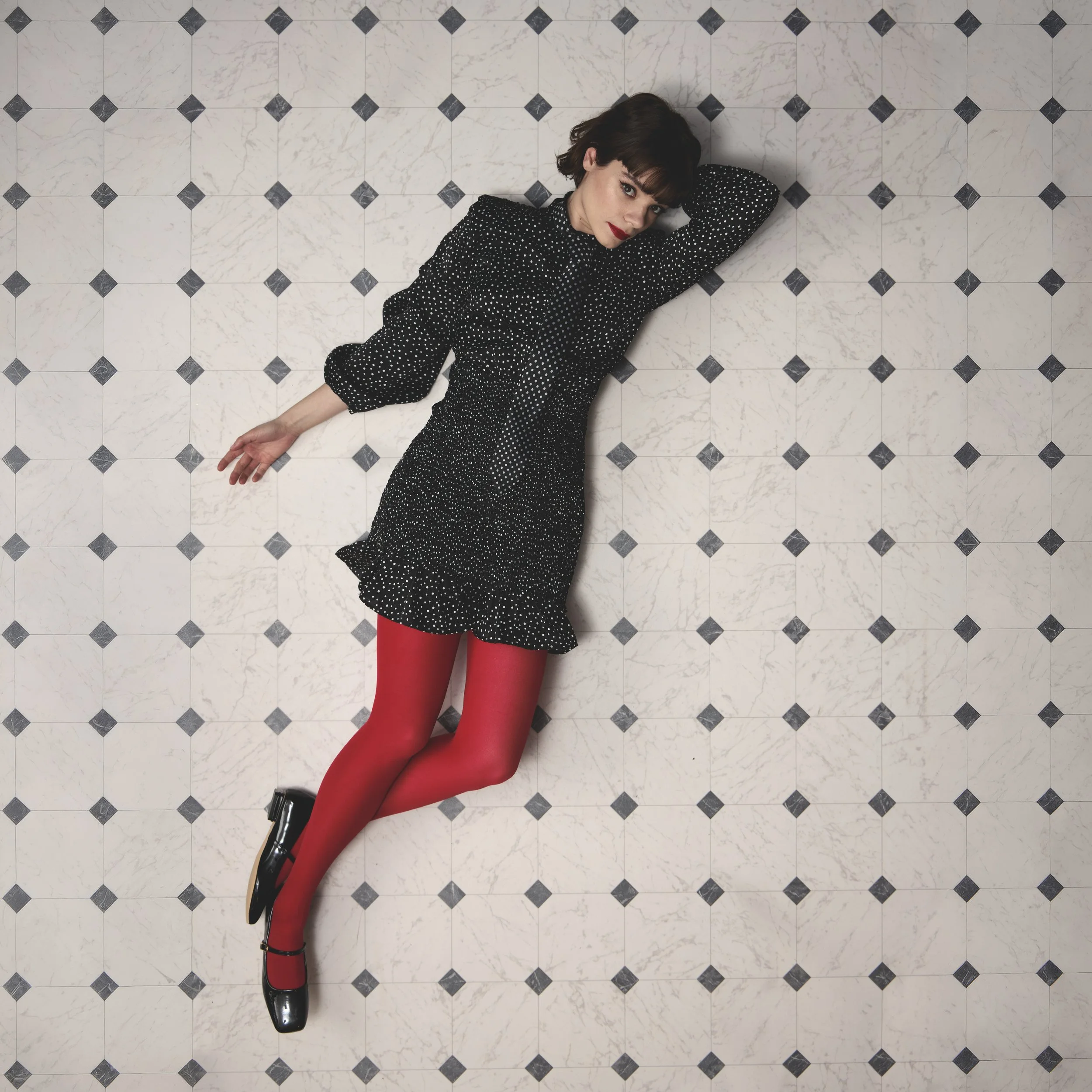 A woman with short dark hair and red lipstick lying on a tiled floor, wearing a black dress with white polka dots, red tights, and black shoes.