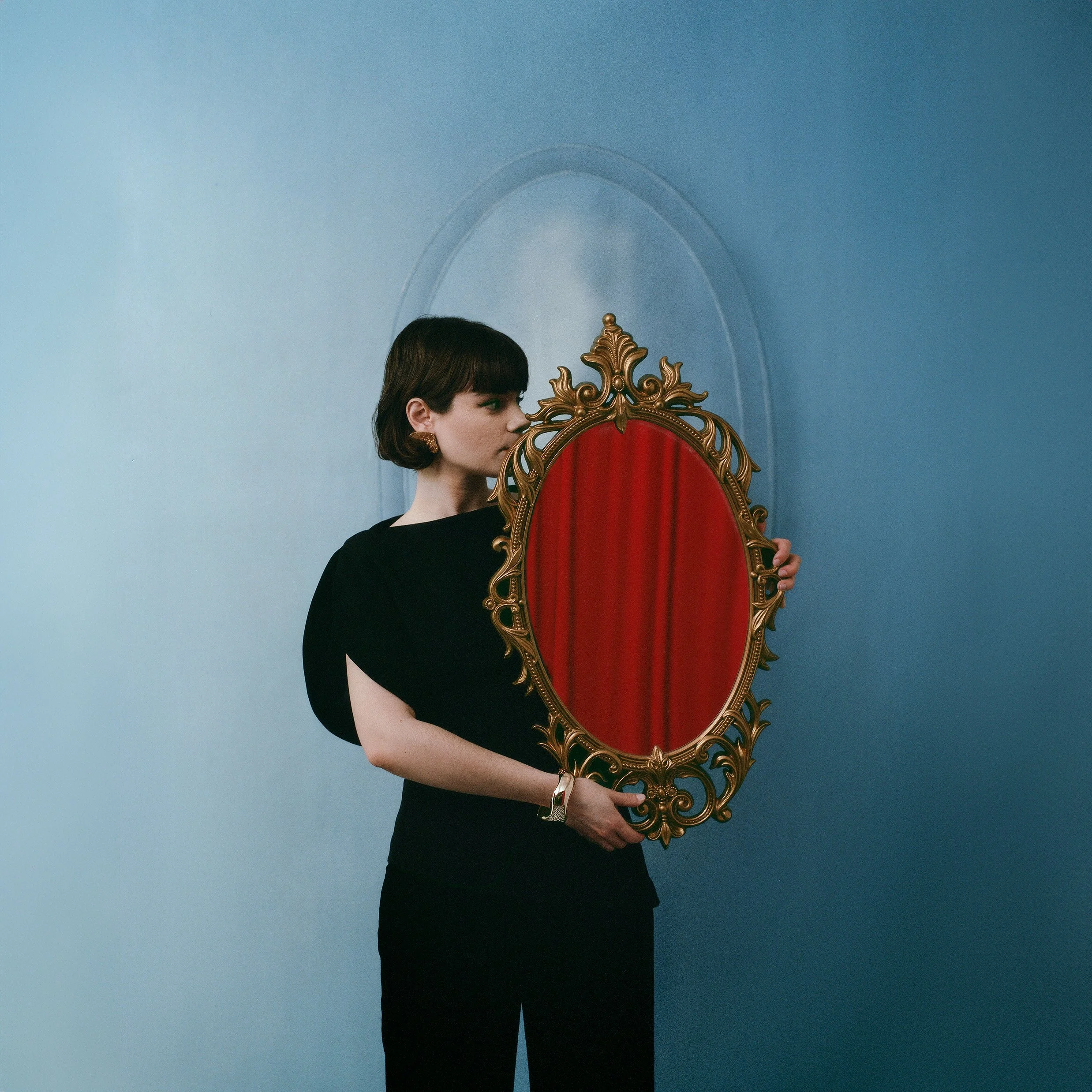 A woman with dark hair and earrings looking at herself in an ornate, gold-framed mirror with red velvet backing, set against a light blue wall.