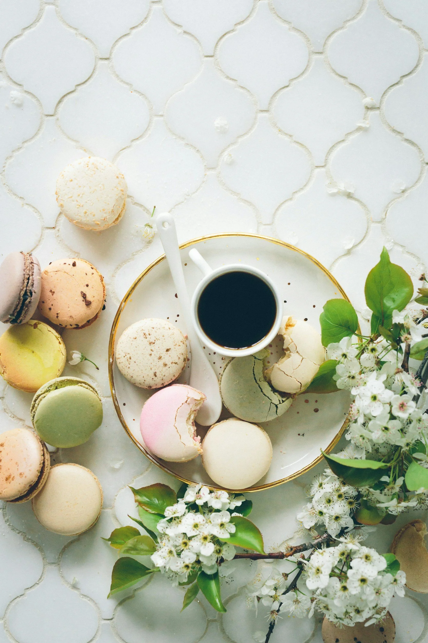 Assorted macarons on a white surface with a cup of black coffee, white flowers, and green leaves.
