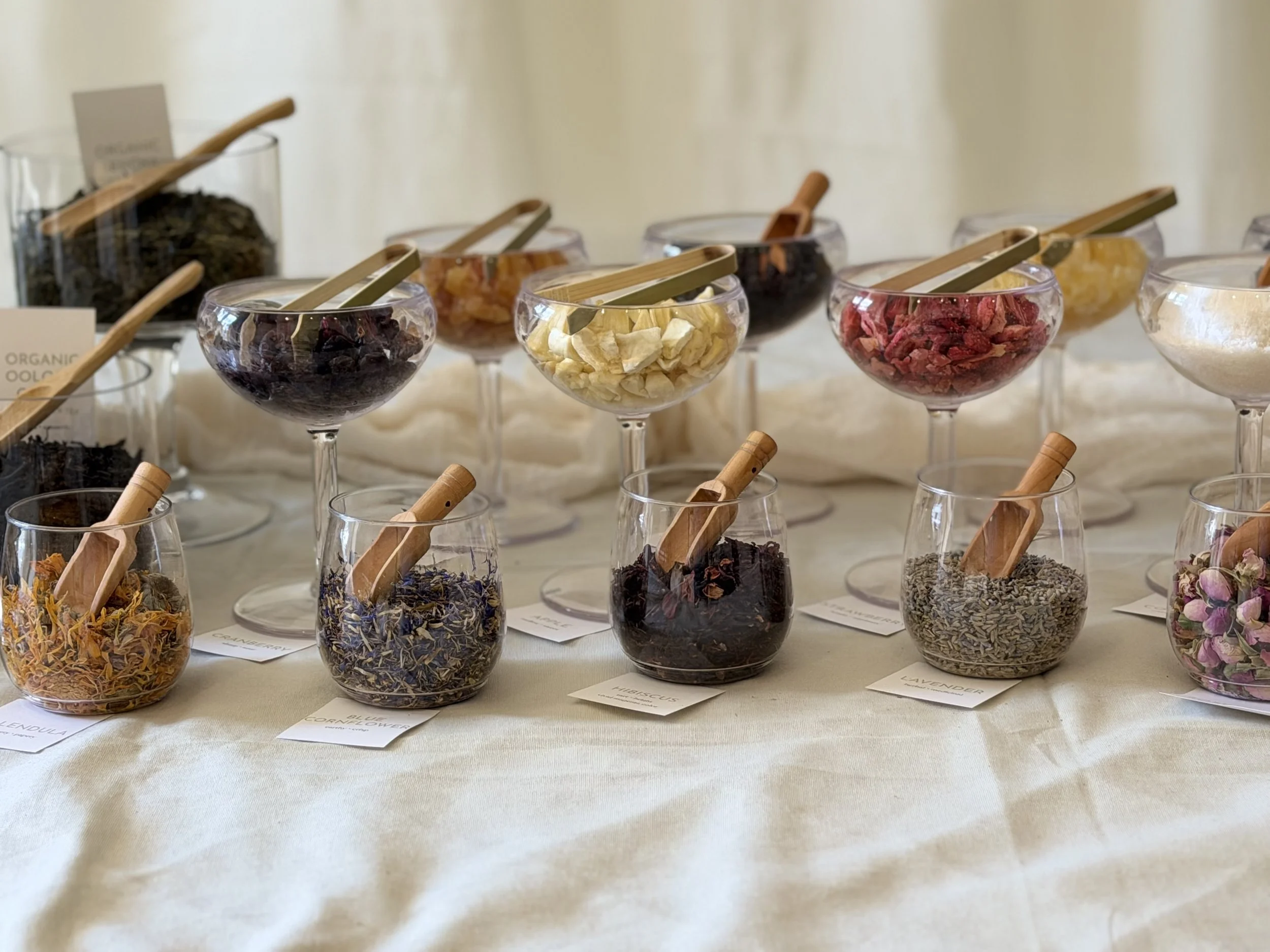 Loose-leaf teas, dried fruits, and edible flowers arranged in glass vessels with scoops and tongs at a tea blending bar.