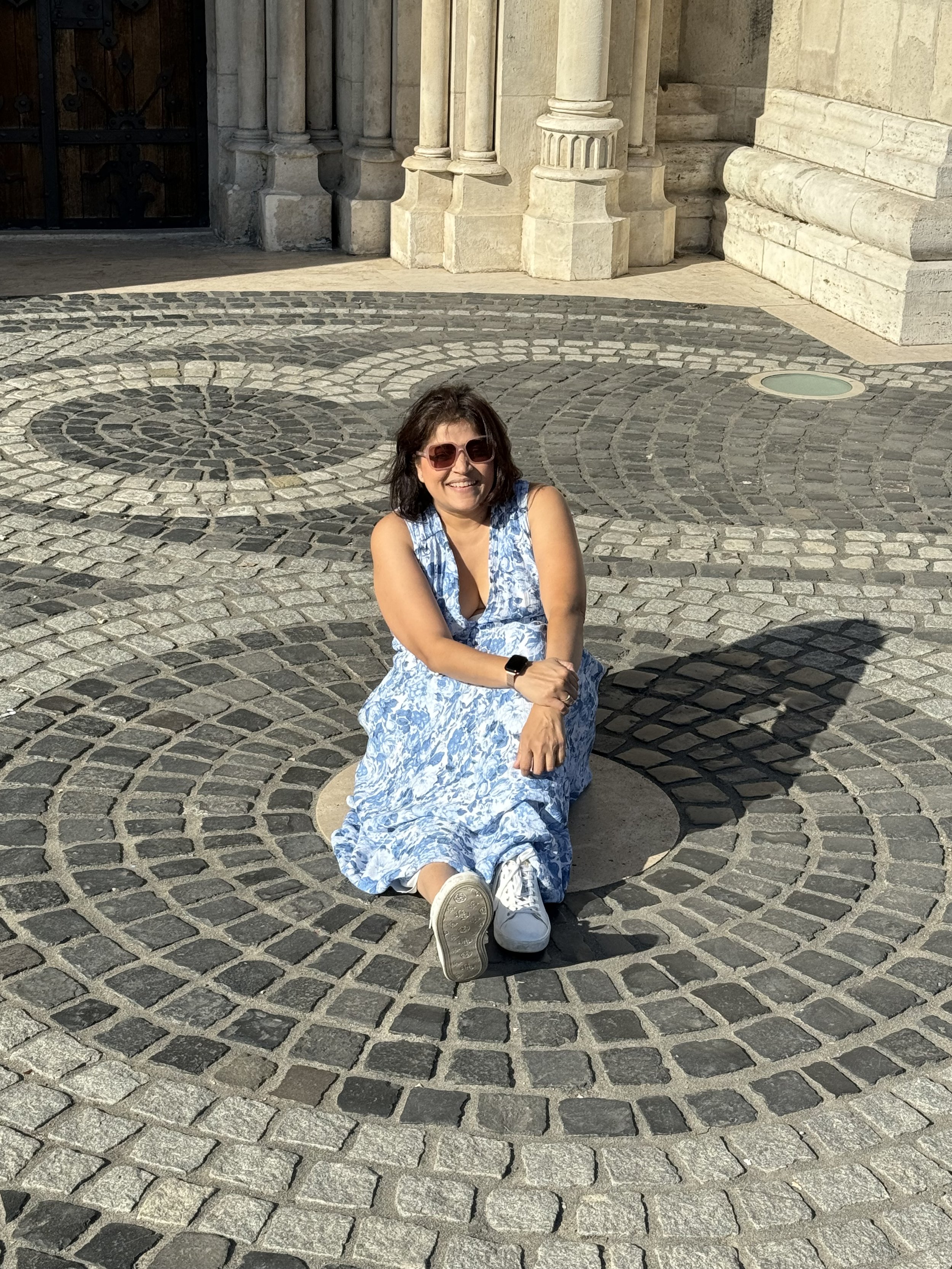 A woman with brown hair wearing sunglasses, a sleeveless blue and white dress, and white sneakers sitting on a circular stone bench on a cobblestone street, smiling at the camera.