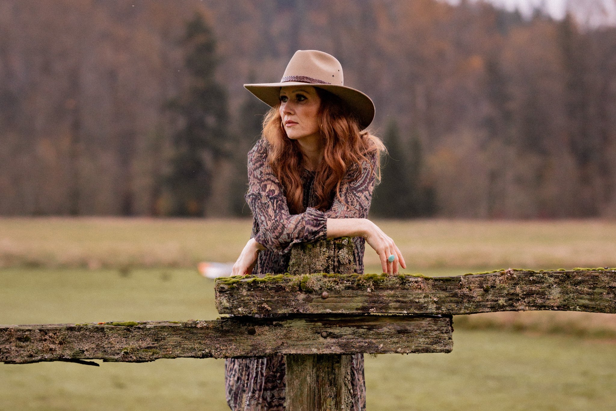 A woman with long red wavy hair wearing a wide-brimmed hat and patterned dress, leaning on a tree stump in an outdoor field during autumn.