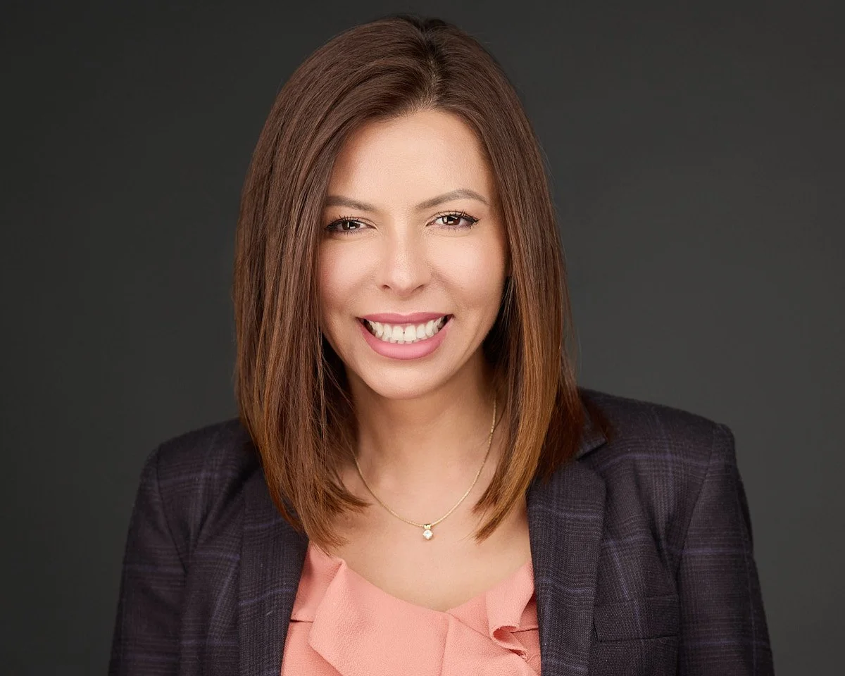 Rocio Vaca, LCSWA woman with shoulder-length light brown hair smiling, wearing a black blazer over a pink blouse, and a delicate gold necklace, against a dark gray background.