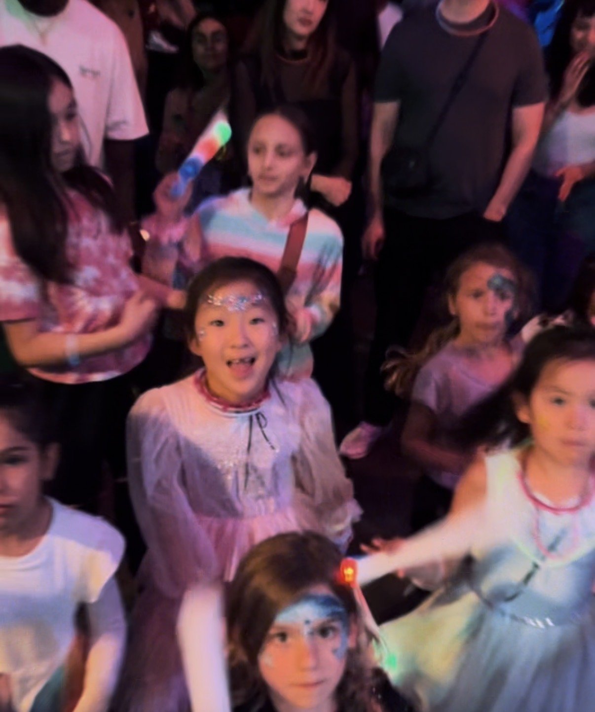 A group of children and teenagers at a lively event, some with face paint, colorful outfits, and glow-in-the-dark accessories, surrounded by adults.