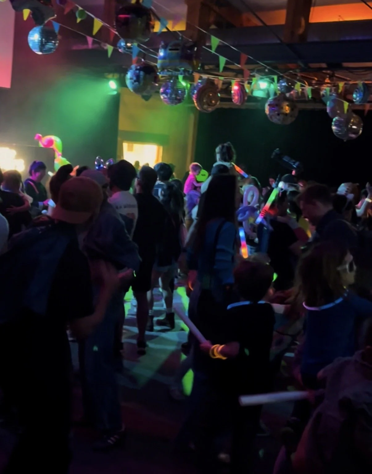 Kids at a party with glow sticks, neon decorations, and disco balls, dancing and socializing under colorful lighting.