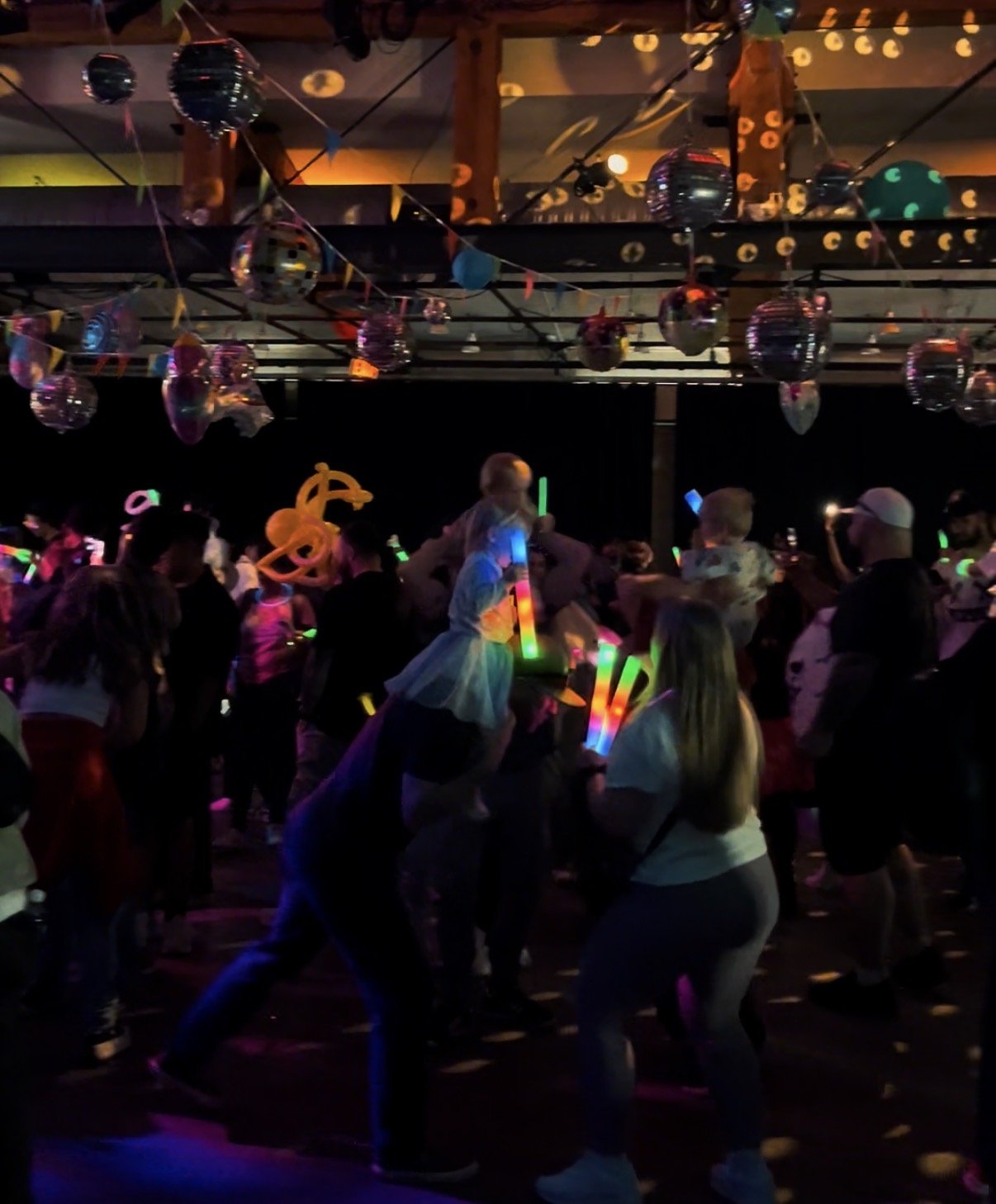 Kids and children dancing at a neon glow party with colorful glow sticks, balloons, and hanging decorations in a dark room.