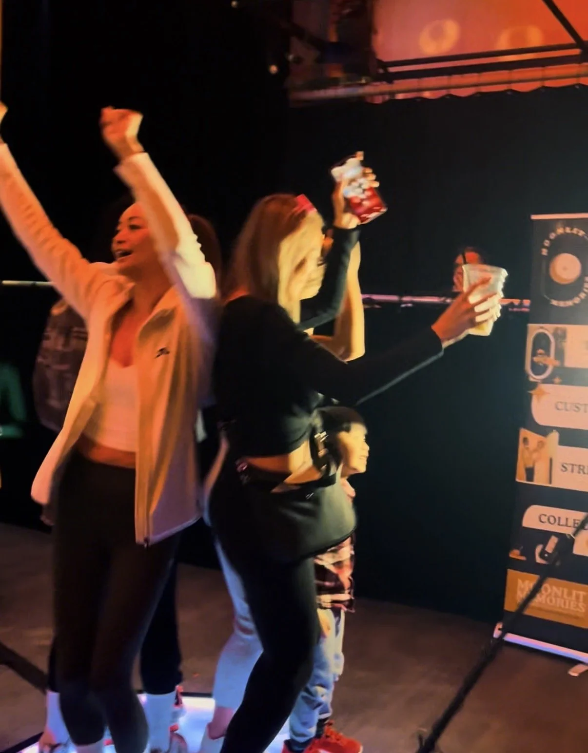 People at a bar or club holding drinks, celebrating, with colorful lighting and a sign with text in the background.