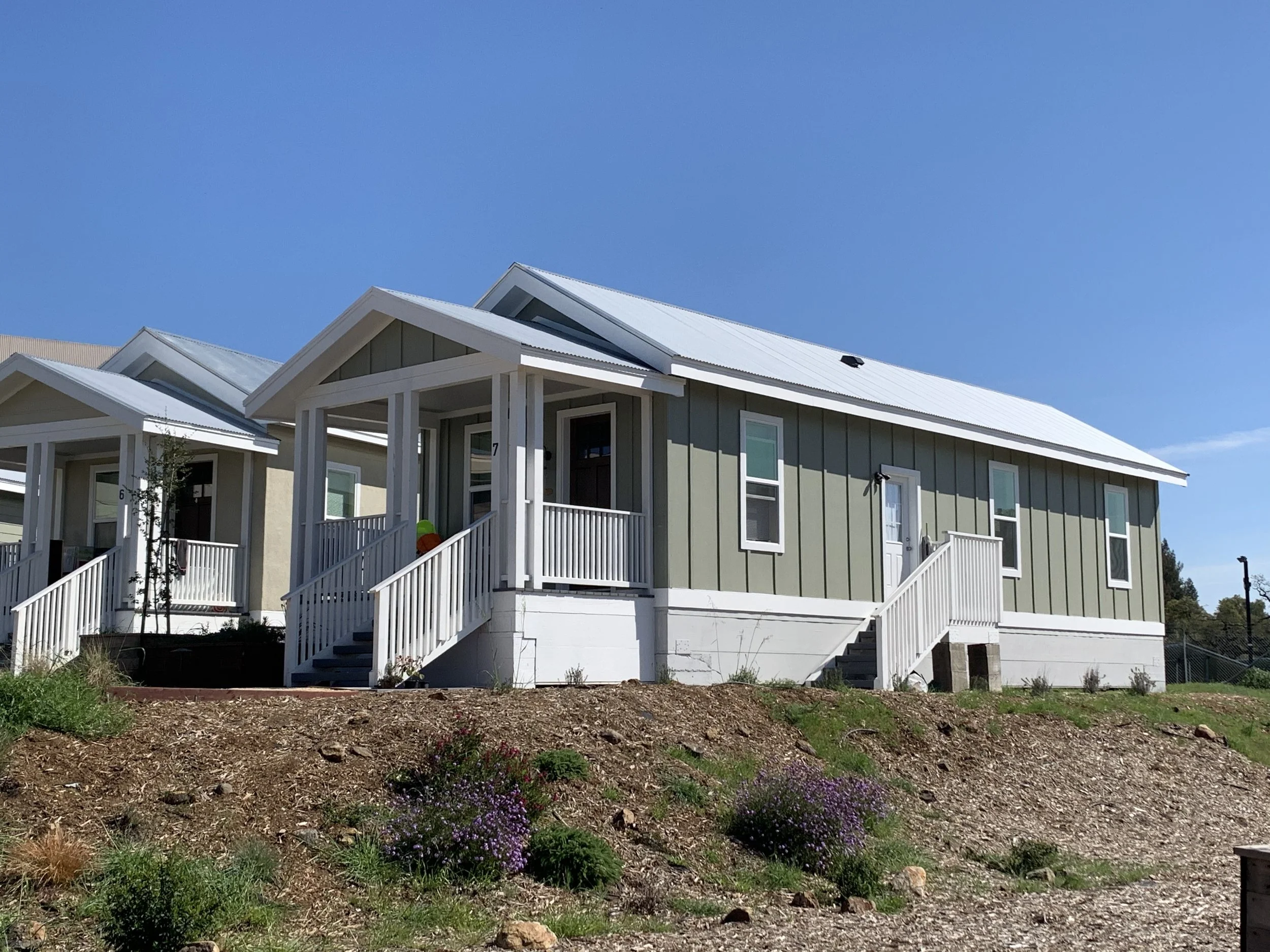 A row of gray houses with white trim and blue skies in the background. The houses have porches with railings and stairs leading up to the entrances. The foreground shows a dirt and grass area with small plants and flowers.