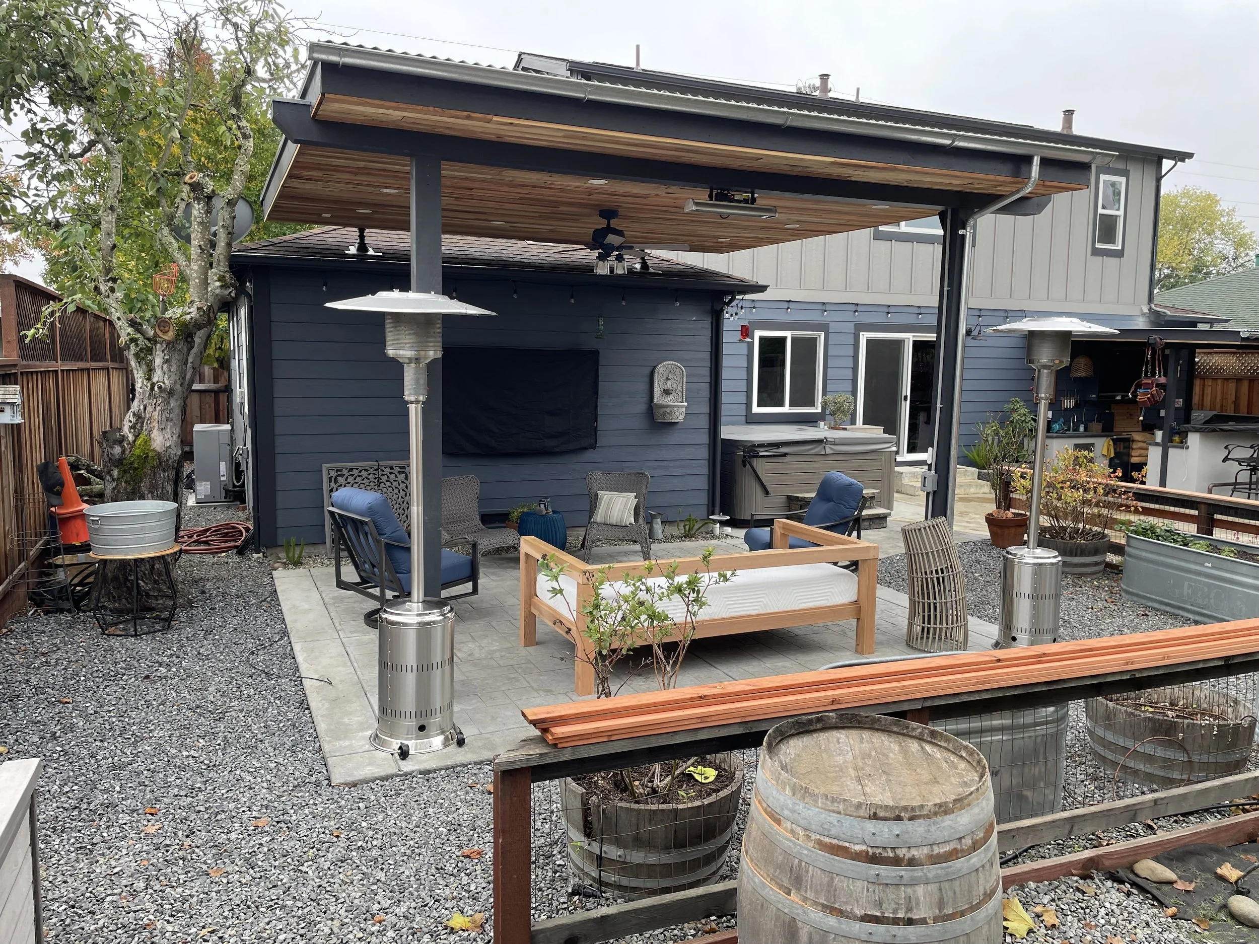 A backyard patio with outdoor furniture including chairs, a table, and tall outdoor heaters. There is a tree with a few leaves, a wooden fence, and a house with a blue exterior. The patio area is paved and surrounded by decorative pots and plants.