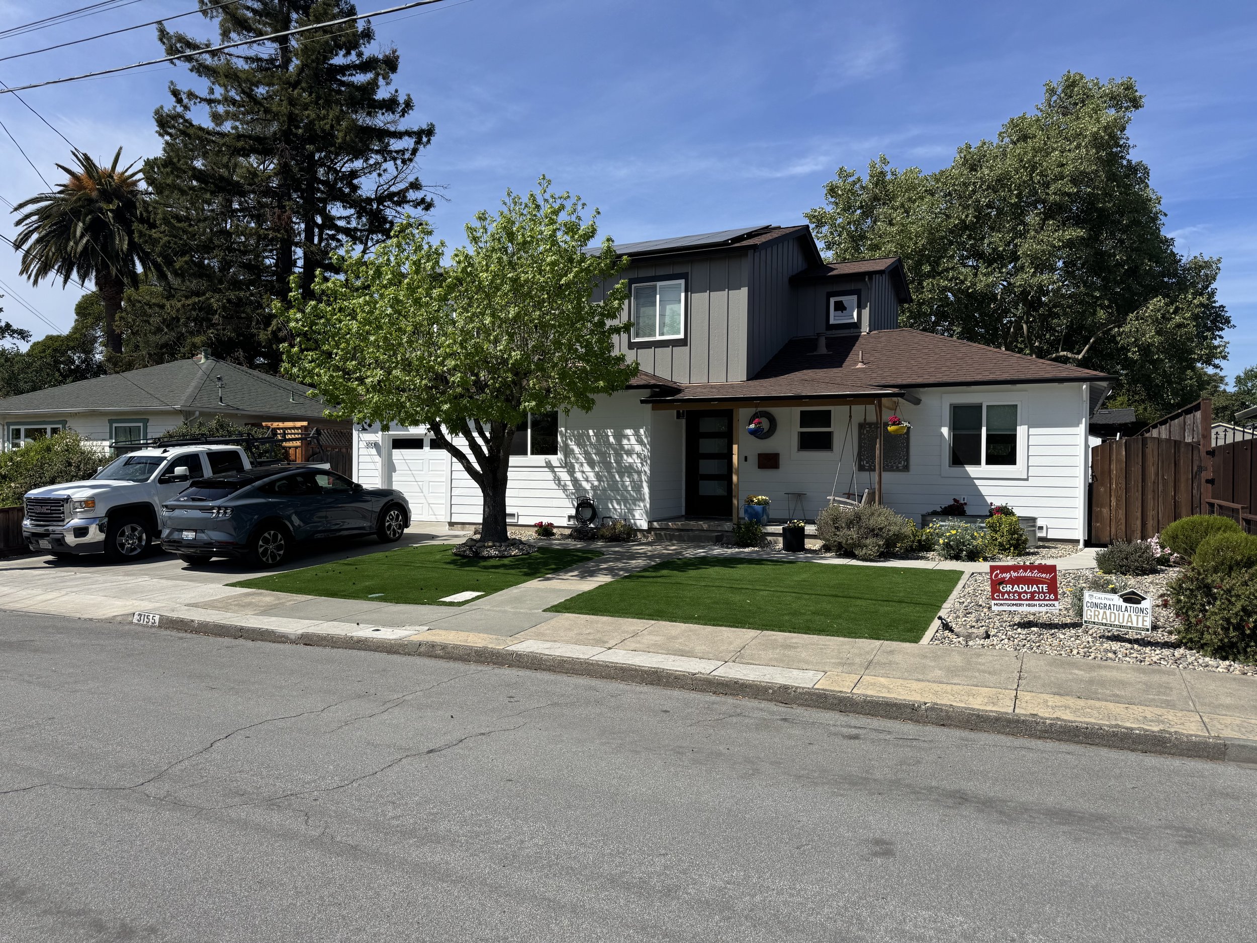 Front view of a two-story house with a gray upper section and white lower section, a small porch with hanging flower baskets, three cars parked in the driveway, and a yard with small plants and flowers. There are two signs on the lawn stating 'Congratulations Graduate of 2026 Montgomery High School.' Tall trees and a house with a dark roof are visible in neighboring properties. The sky is mostly clear with some clouds.