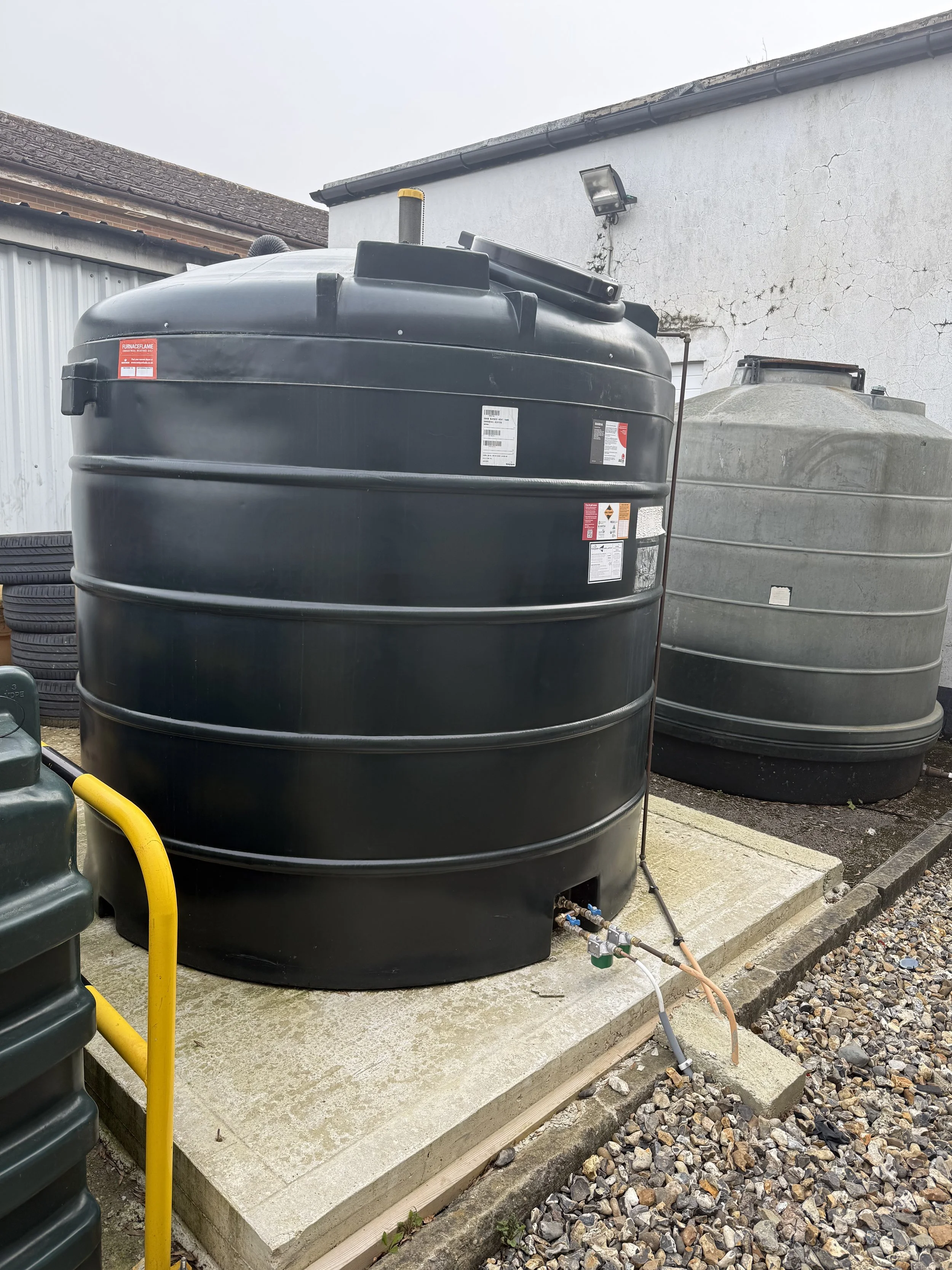 Two large tanks outdoors, one black and one gray, connected to a small pipe and valve, with a concrete pad and gravel ground surrounding them.