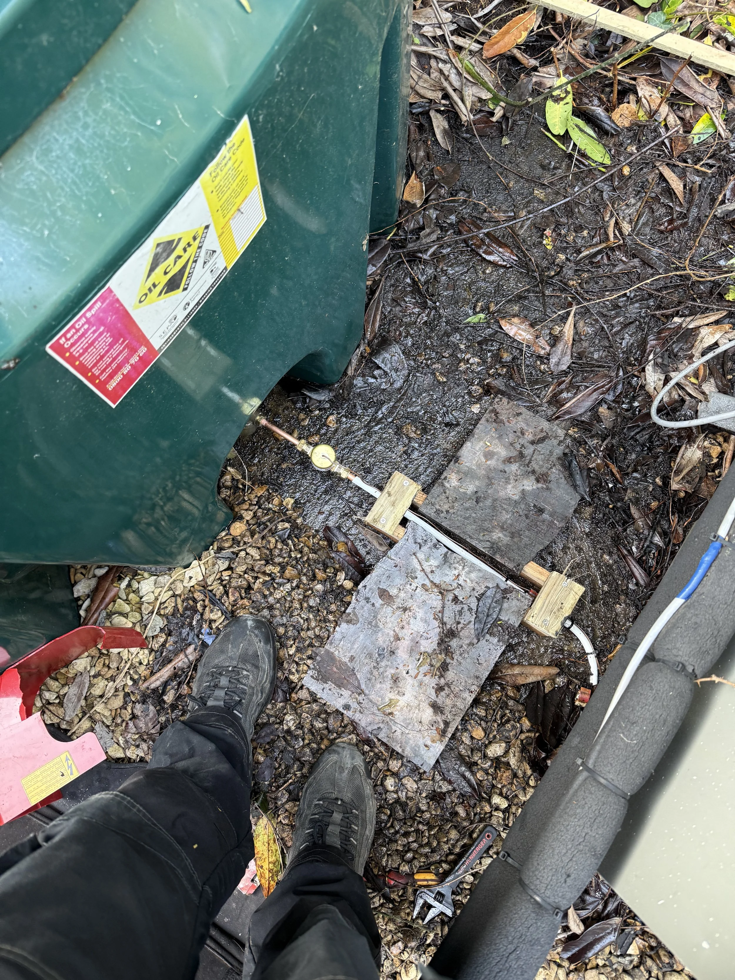 View of a person's shoes standing on gravel and wet ground near a green utility box with electrical cables, a wrench, and a metal plate.