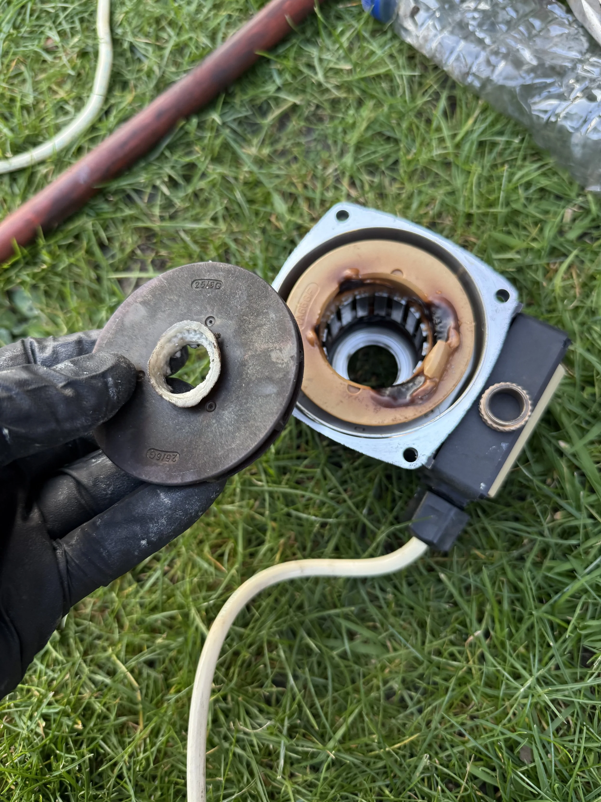 Person wearing black gloves holding a worn mechanical part over an open bearing housing, which contains brown grease and internal components, on green grass.