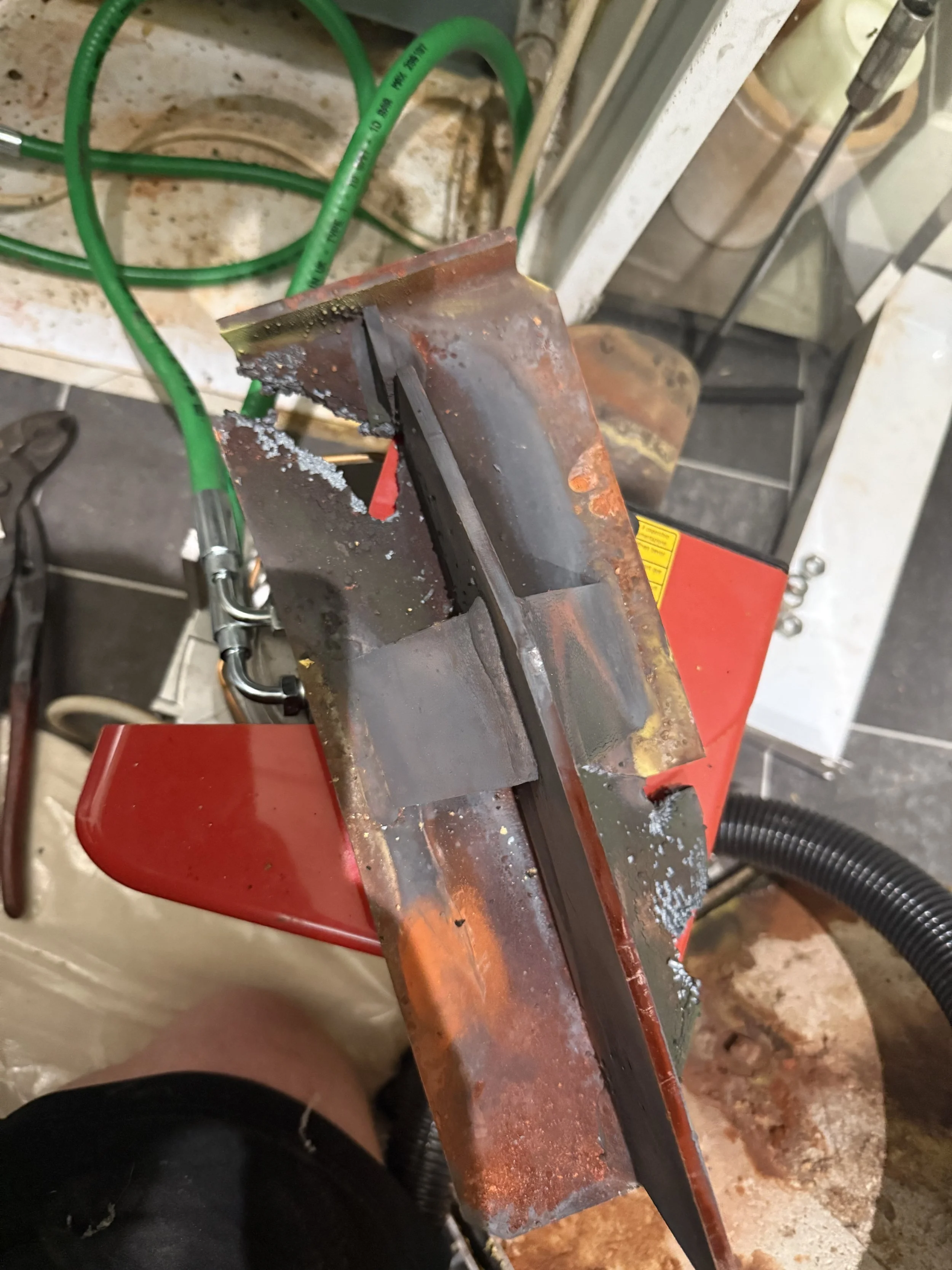 Close-up of a metal piece with rust and weld marks on a workbench with green garden hoses and tools in the background.
