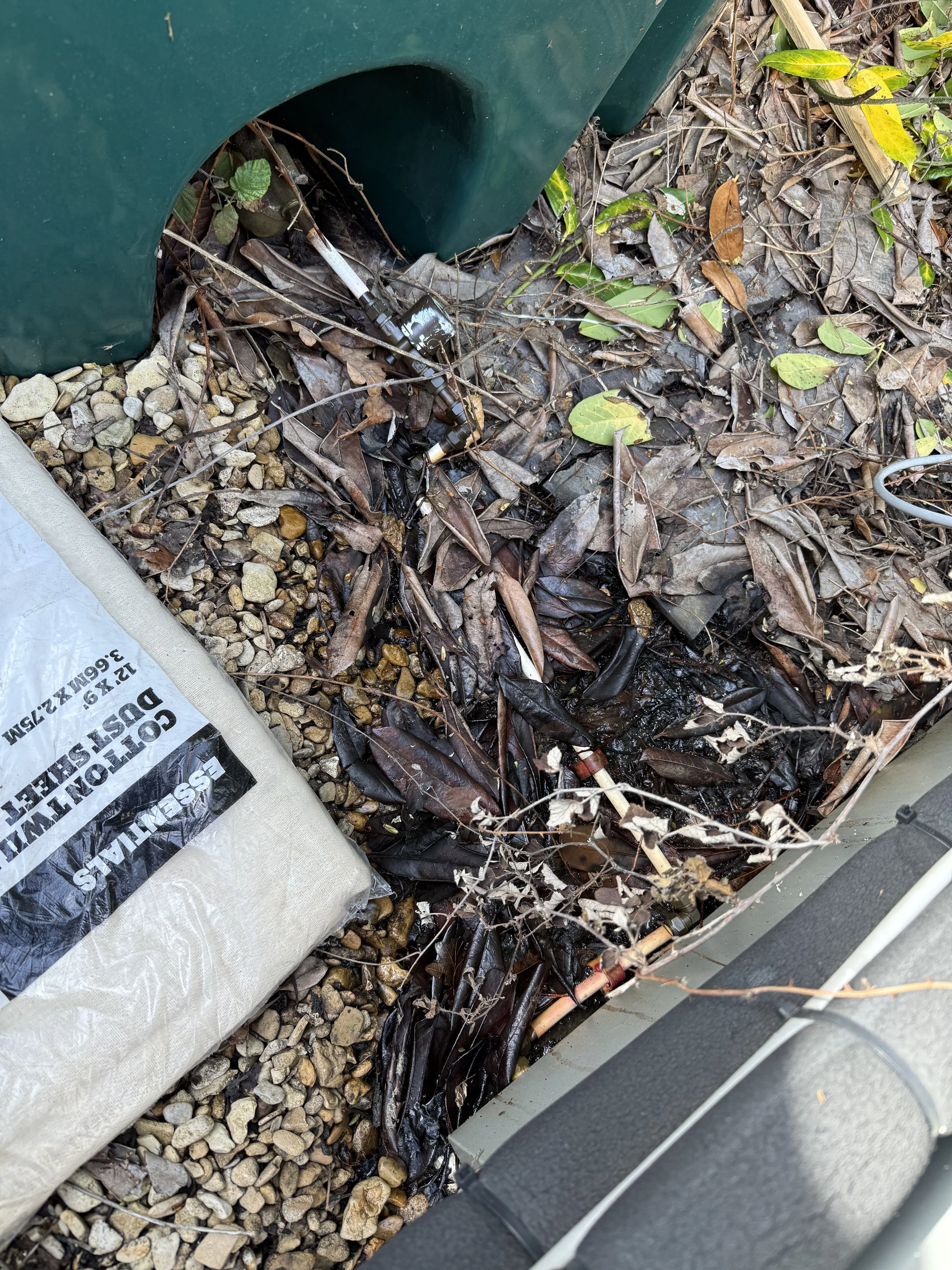 Pipeline leak with water pooling on the ground surrounded by dead leaves and rocks, with a bag labeled 'GOTTLIEB'S DUSTLESS' on the side.
