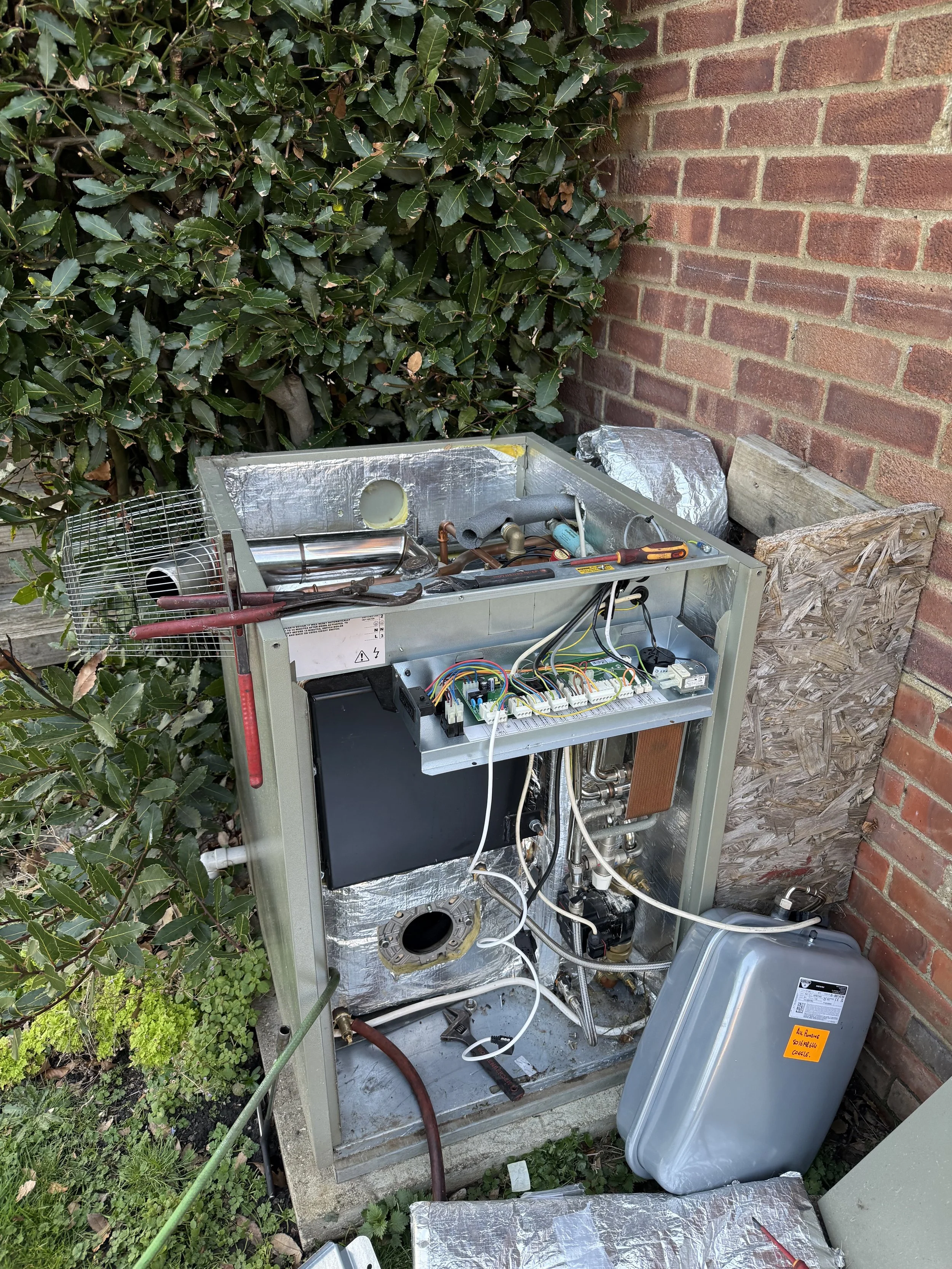 Open HVAC unit with exposed internal components and wiring outdoors near a brick wall and green shrubbery.