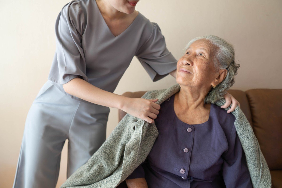 A caregiver assisting an elderly woman with a warm smile in a cozy room.