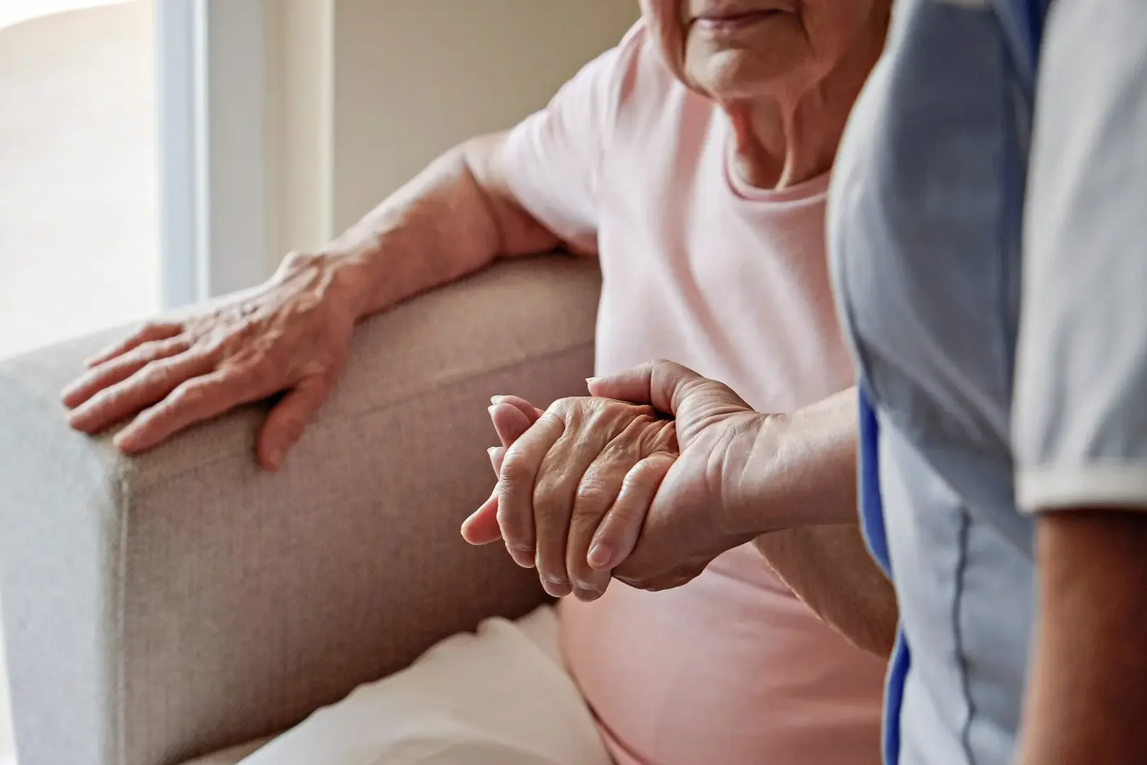 A close-up of an elderly woman and a caregiver holding hands, with the elderly woman resting her arm on a beige couch.