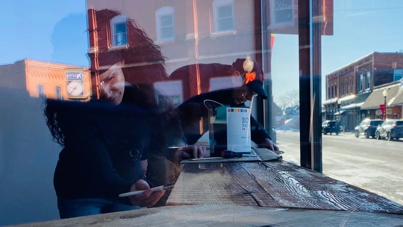 People sitting at a wooden table inside a cafe, seen through a large glass window, with a street scene outside including buildings and parked cars.