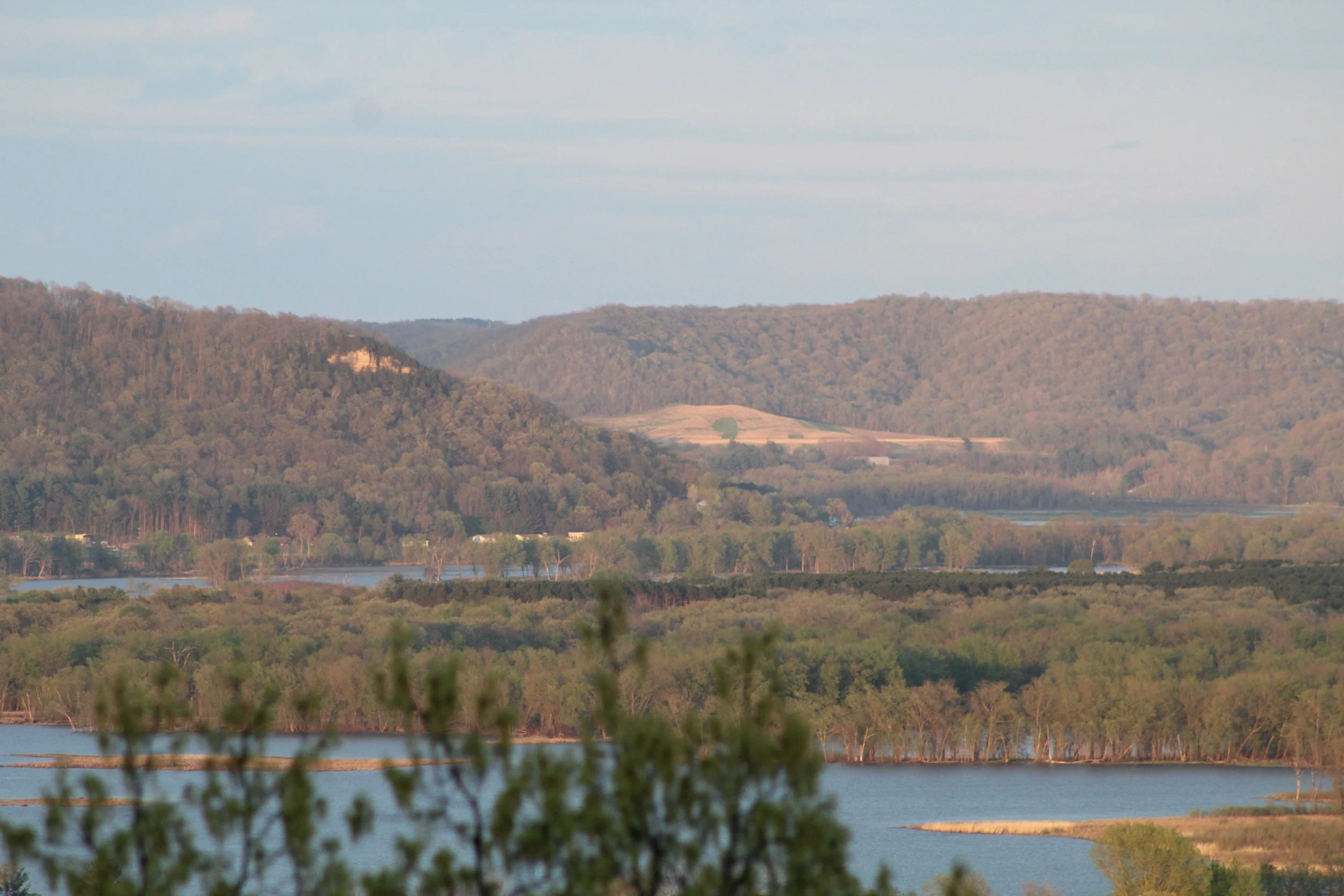 A landscape view of rolling hills covered in trees with a river or lake in the foreground, under a partly cloudy sky.